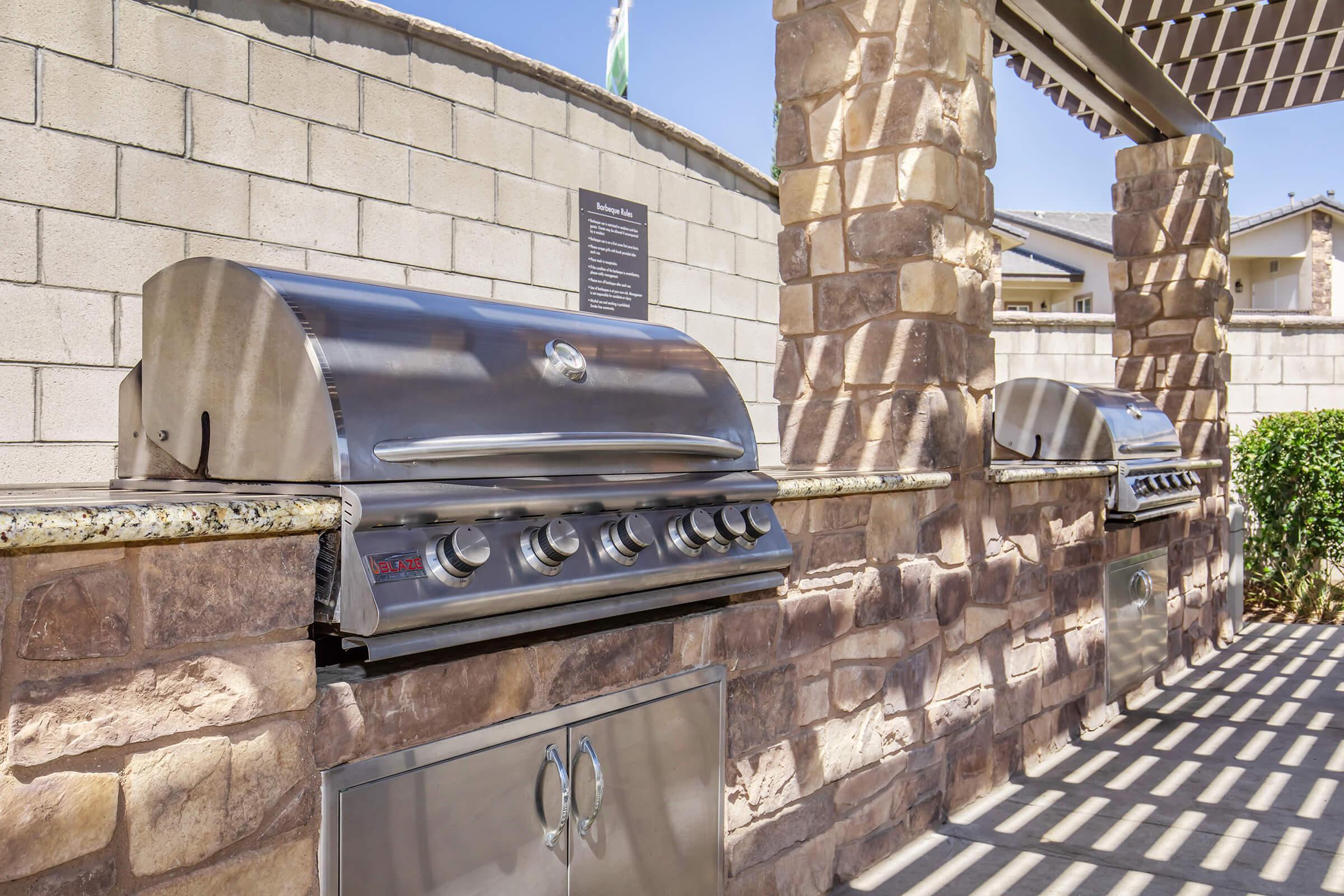 Outdoor grilling area featuring three stainless steel gas grills on stone countertops. The grills are set against a textured stone wall, with a pergola structure providing shade overhead. Greenery is visible in the background, enhancing the inviting atmosphere for outdoor cooking and dining.