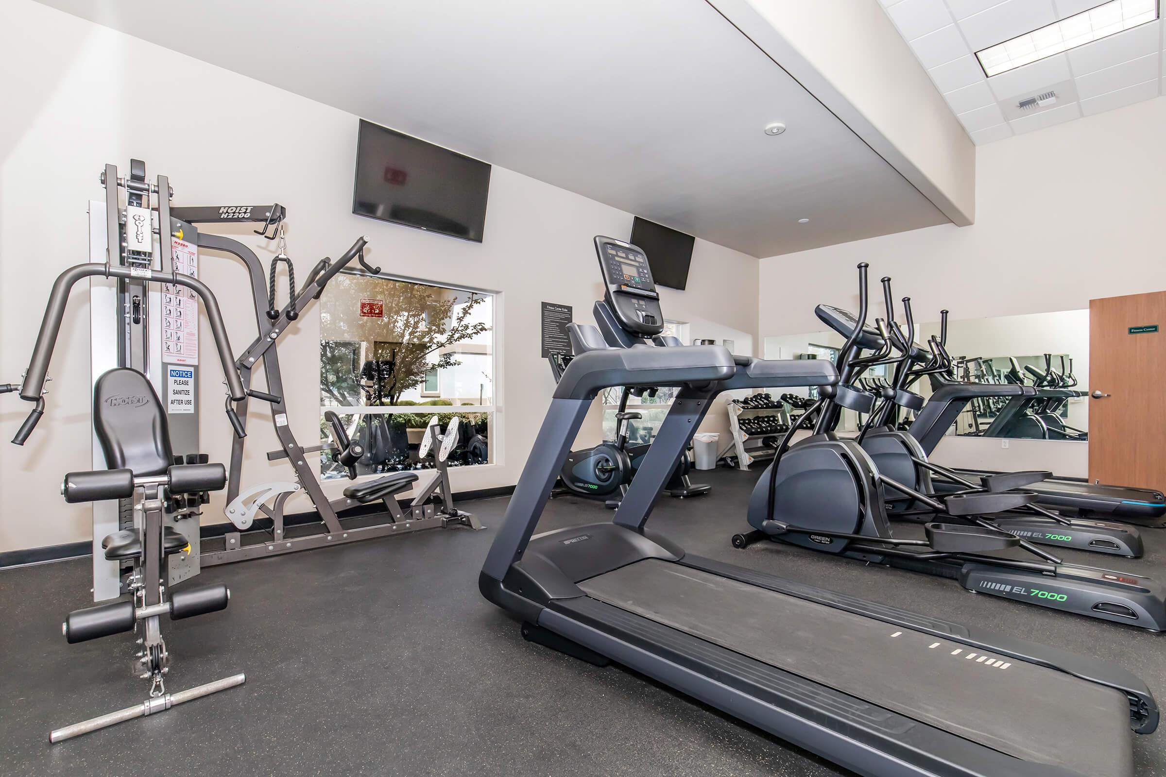 A well-equipped gym featuring various exercise machines, including treadmills, an elliptical trainer, and a multi-station weight machine. The room has large mirrors, flat-screen TVs, and a clean, modern design, providing a bright and inviting space for workouts.