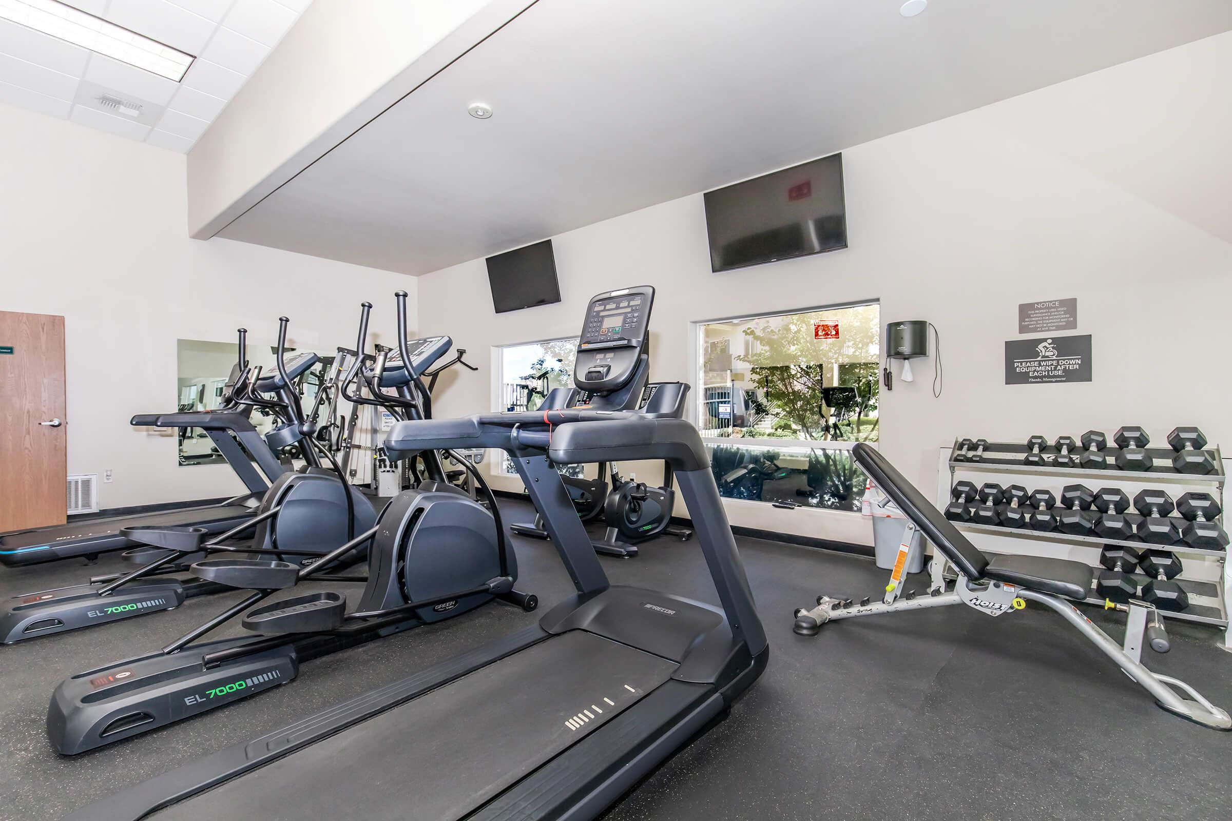 A fitness room featuring several exercise machines including treadmills, stationary bikes, and a weight bench. There are also free weights arranged neatly on a rack. Large mirrors reflect the equipment, and two screens are mounted on the wall for entertainment or instruction.