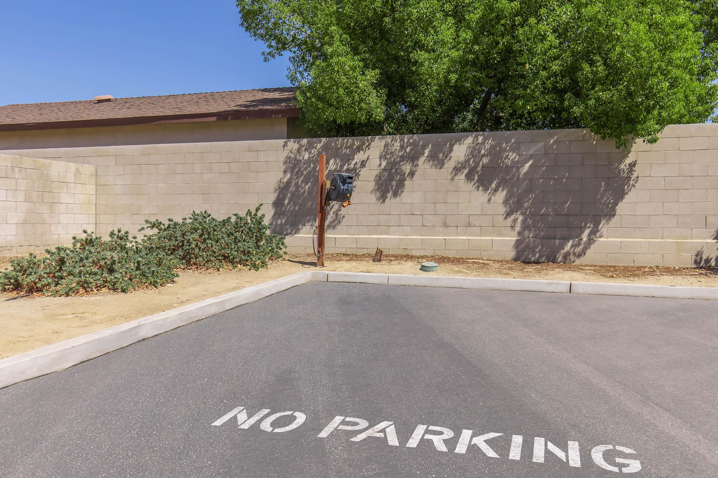 A paved area marked with "NO PARKING" in white paint. A nearby wall provides a backdrop, and there is a mailbox attached to a wooden post on the left side of the image. Sparse vegetation is present on the ground, with a clear blue sky above.