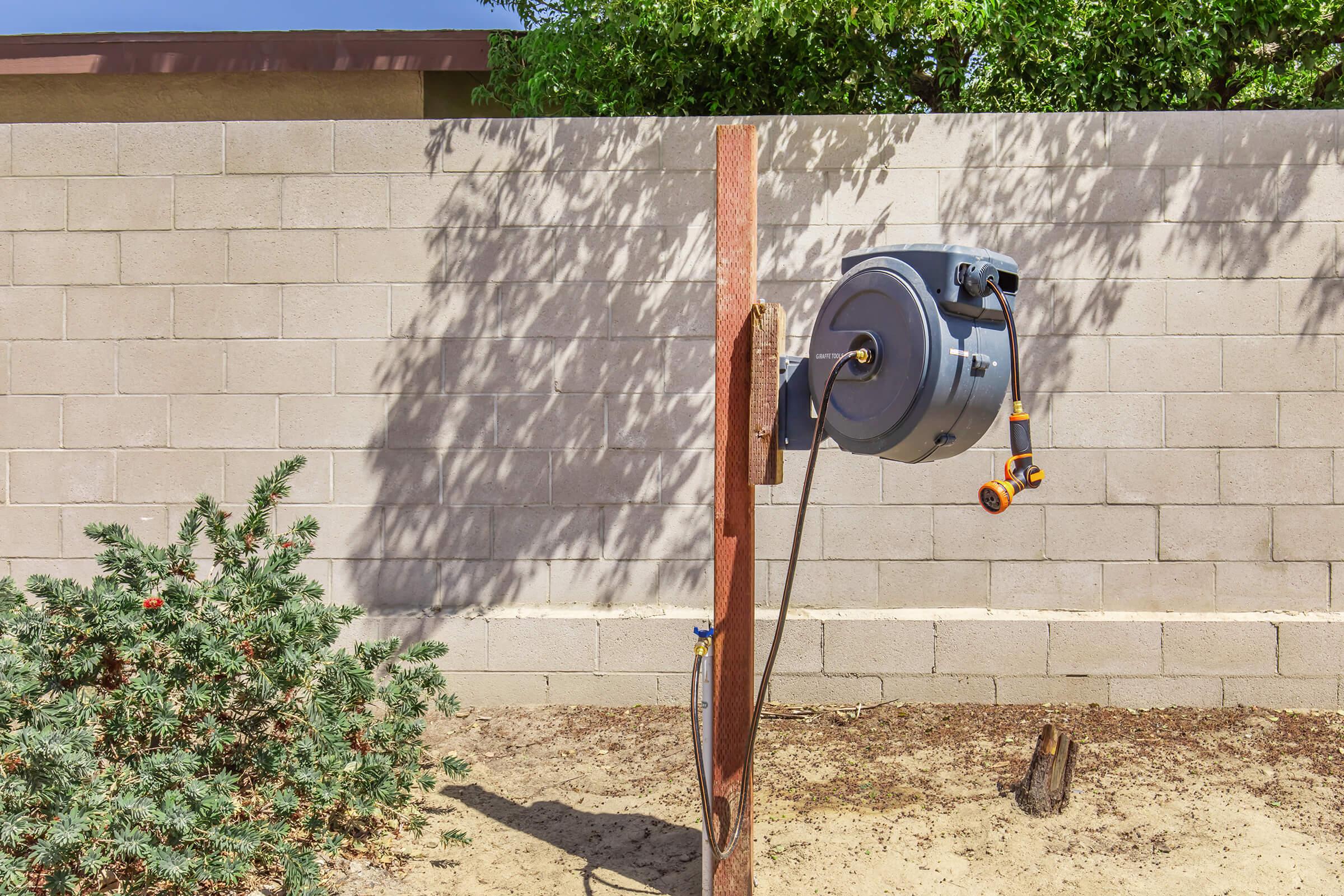 A wall-mounted hose reel attached to a wooden post in a sandy yard, with a bush nearby and a gray block wall in the background. Shadows from the surrounding greenery create a dappled effect on the wall.