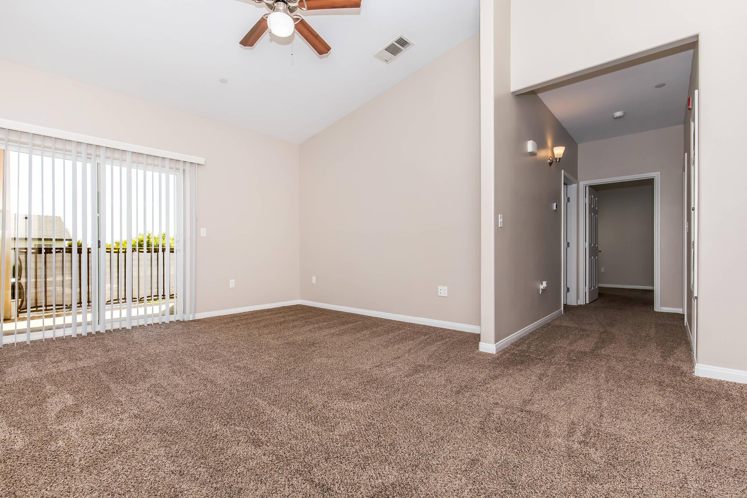 Spacious interior of a light-colored room with a high ceiling, beige walls, and plush carpeting. There's a ceiling fan and sliding glass doors leading to a balcony. A doorway to another room is visible in the background, creating an inviting and open atmosphere.