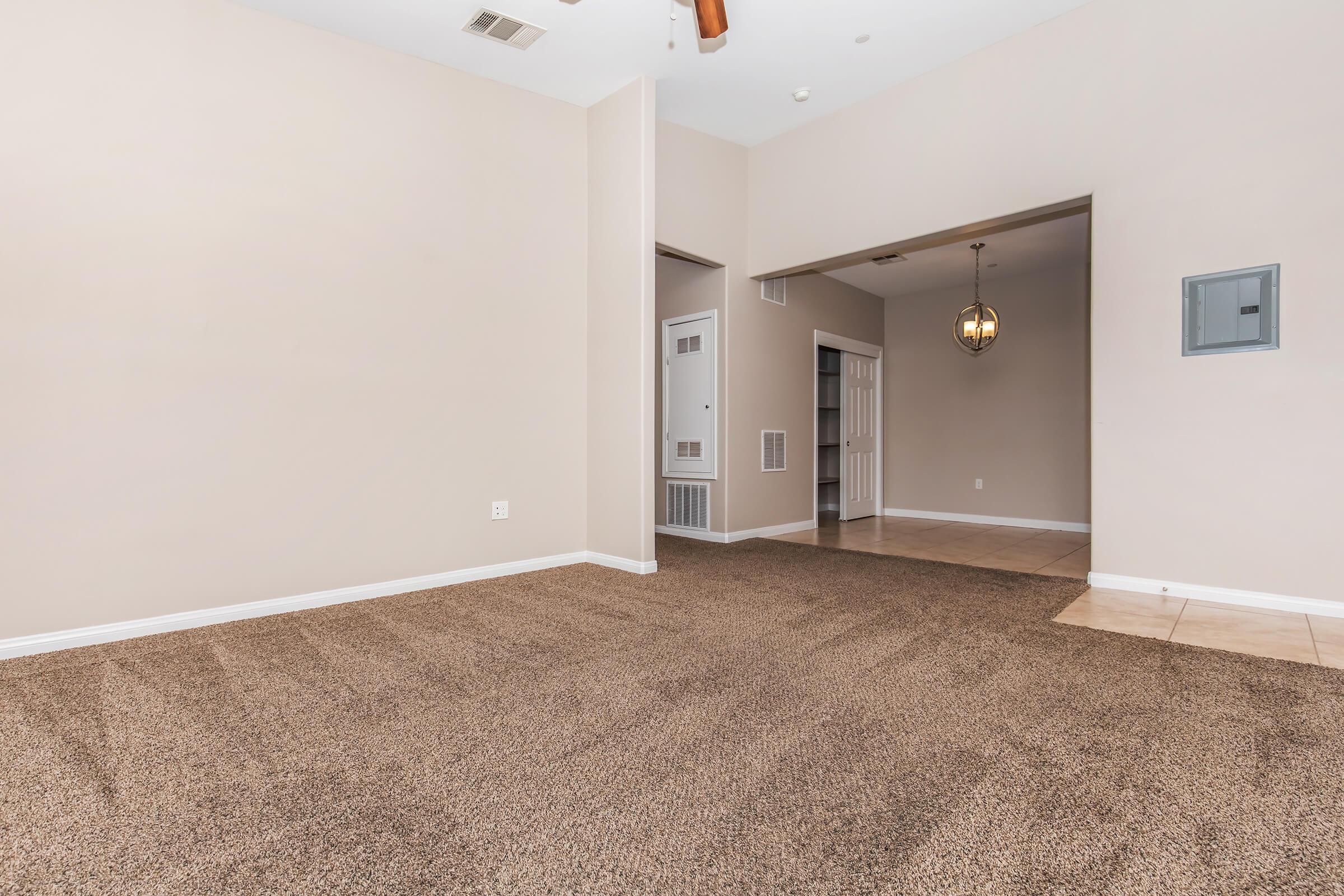 A spacious living area with light beige walls and brown carpet. The room features an archway leading to another space, a pendant light fixture hanging from the ceiling, and a door on the left leading outside. The floor transitions to tile towards the back, creating a cozy and open atmosphere.