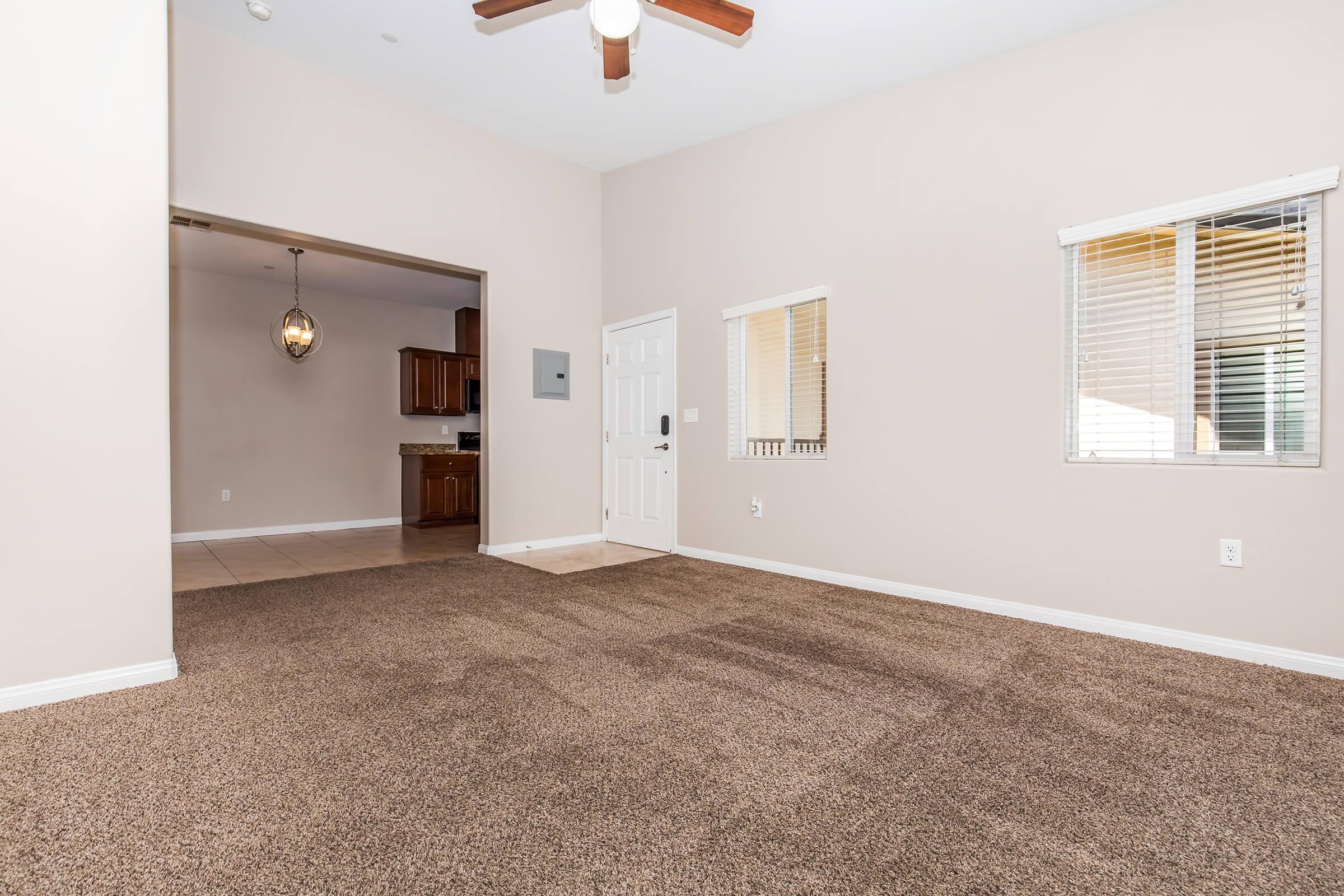A spacious living room with beige walls and carpet flooring. A ceiling fan is mounted above. To the left, there’s an archway leading to a kitchen area with wooden cabinets visible. Natural light comes through two windows, and a door leads to another room, creating an open and inviting atmosphere.