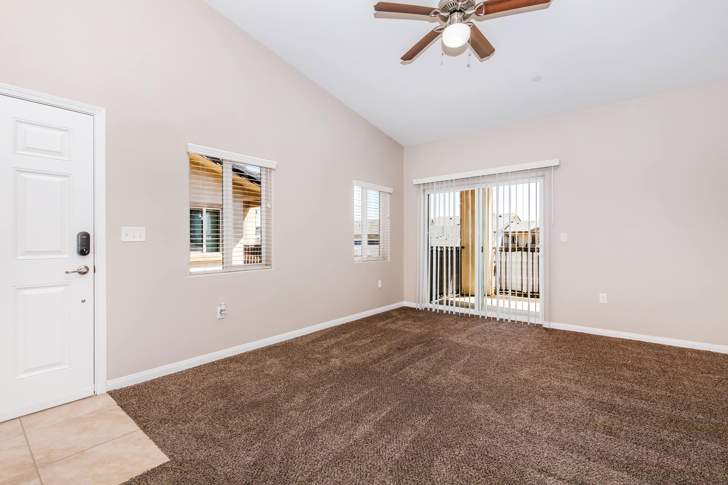 A spacious, empty living room featuring light beige walls and carpet flooring. It has a ceiling fan, two small windows, and a sliding glass door that leads to a balcony. Natural light fills the room, creating an inviting atmosphere.