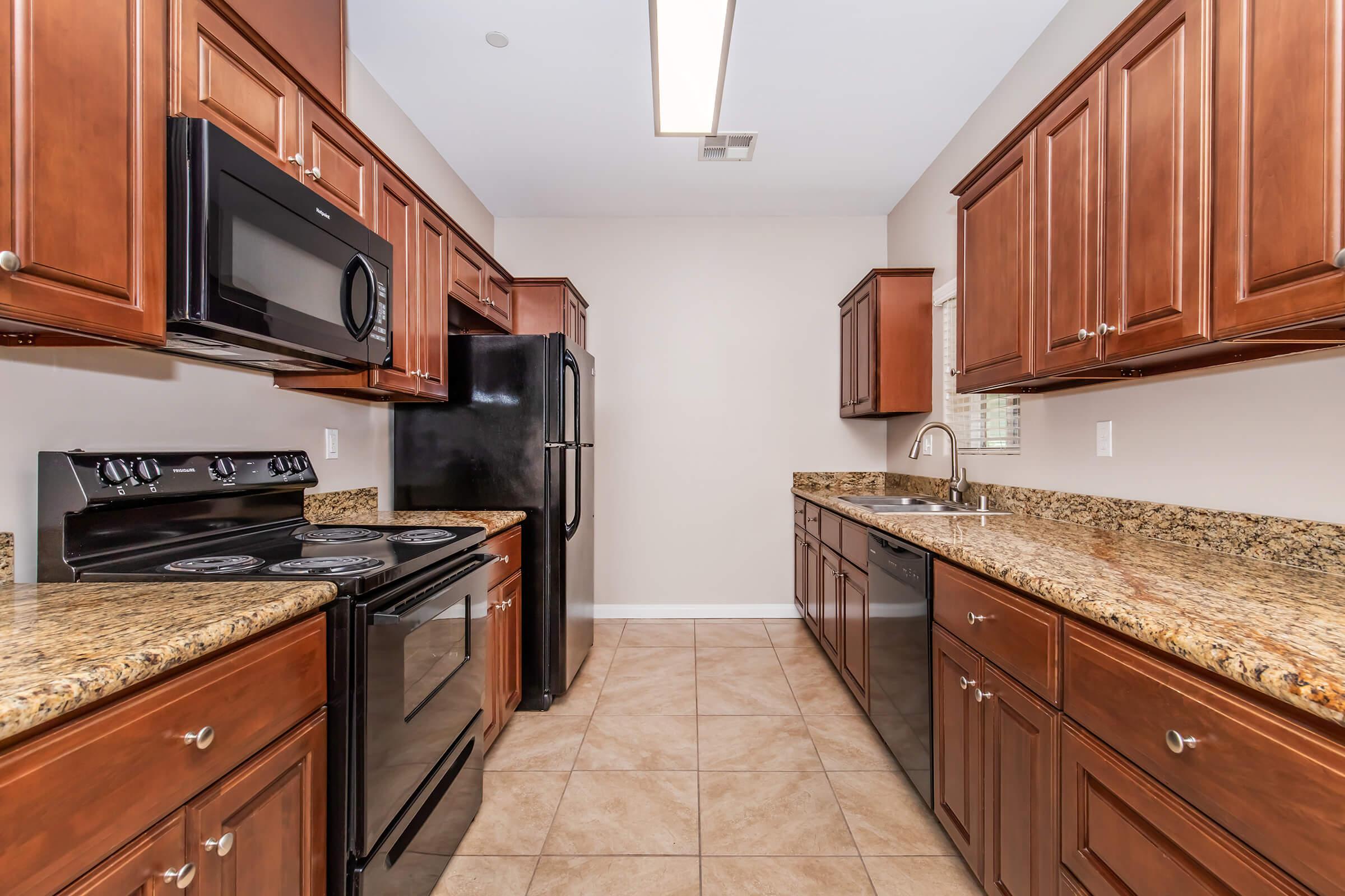 Modern kitchen featuring dark wood cabinets, granite countertops, and stainless steel appliances. The space includes a black oven, microwave, and refrigerator, along with a sink. The floor is tiled, and the walls are painted a light color, creating a bright and airy atmosphere.
