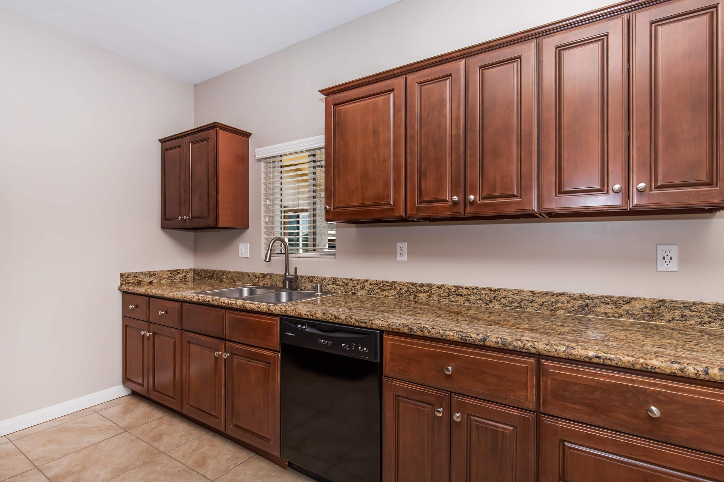 A modern kitchen featuring dark wood cabinets, a granite countertop, a stainless steel sink, and a black dishwasher. The walls are painted a light beige, and there is a window with blinds allowing natural light into the space. The floor is tiled.