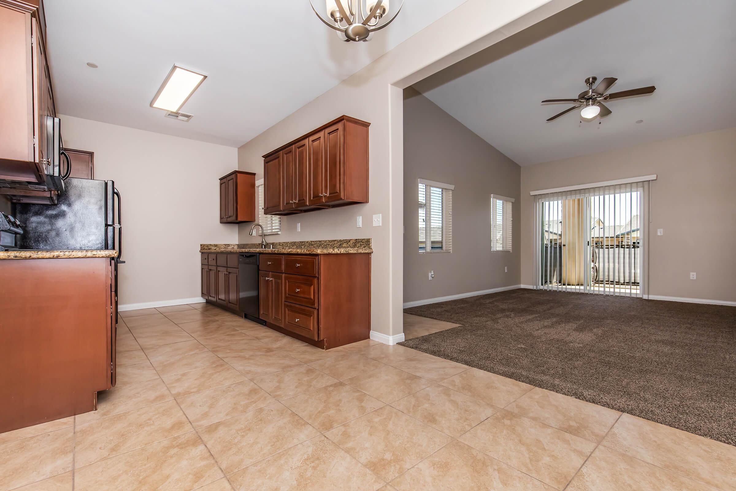 A spacious kitchen with dark wooden cabinets, granite countertops, and stainless steel appliances is visible. The floor is tiled, leading into a carpeted living area with high ceilings and a ceiling fan. Large windows allow natural light, and a sliding door opens to a patio area.