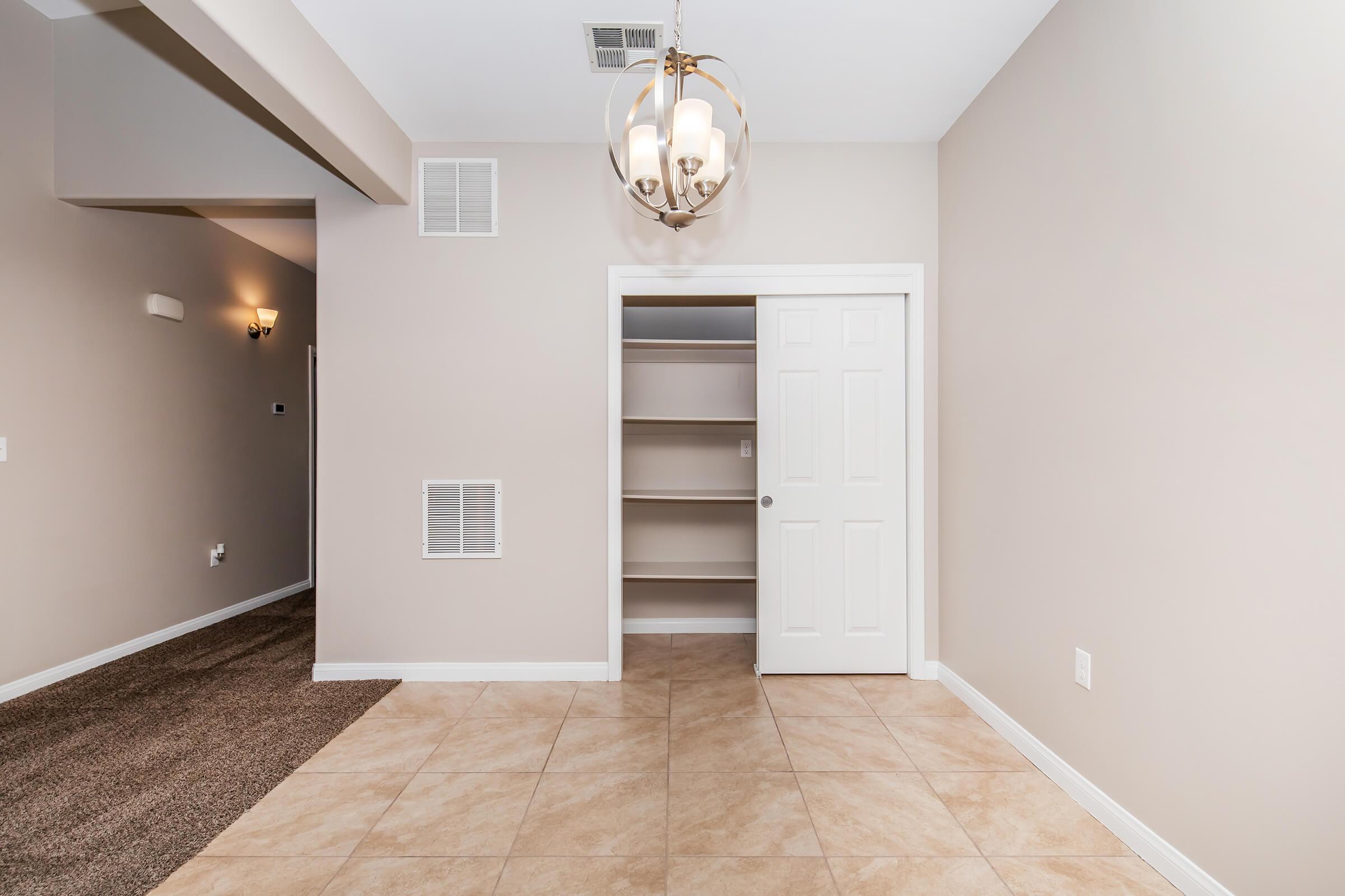 A well-lit room with neutral-colored walls and tile flooring. A closet with white doors is visible on the right, featuring open shelves inside. A decorative chandelier hangs from the ceiling, and there is a textured wall, possibly leading to another room, in the background.