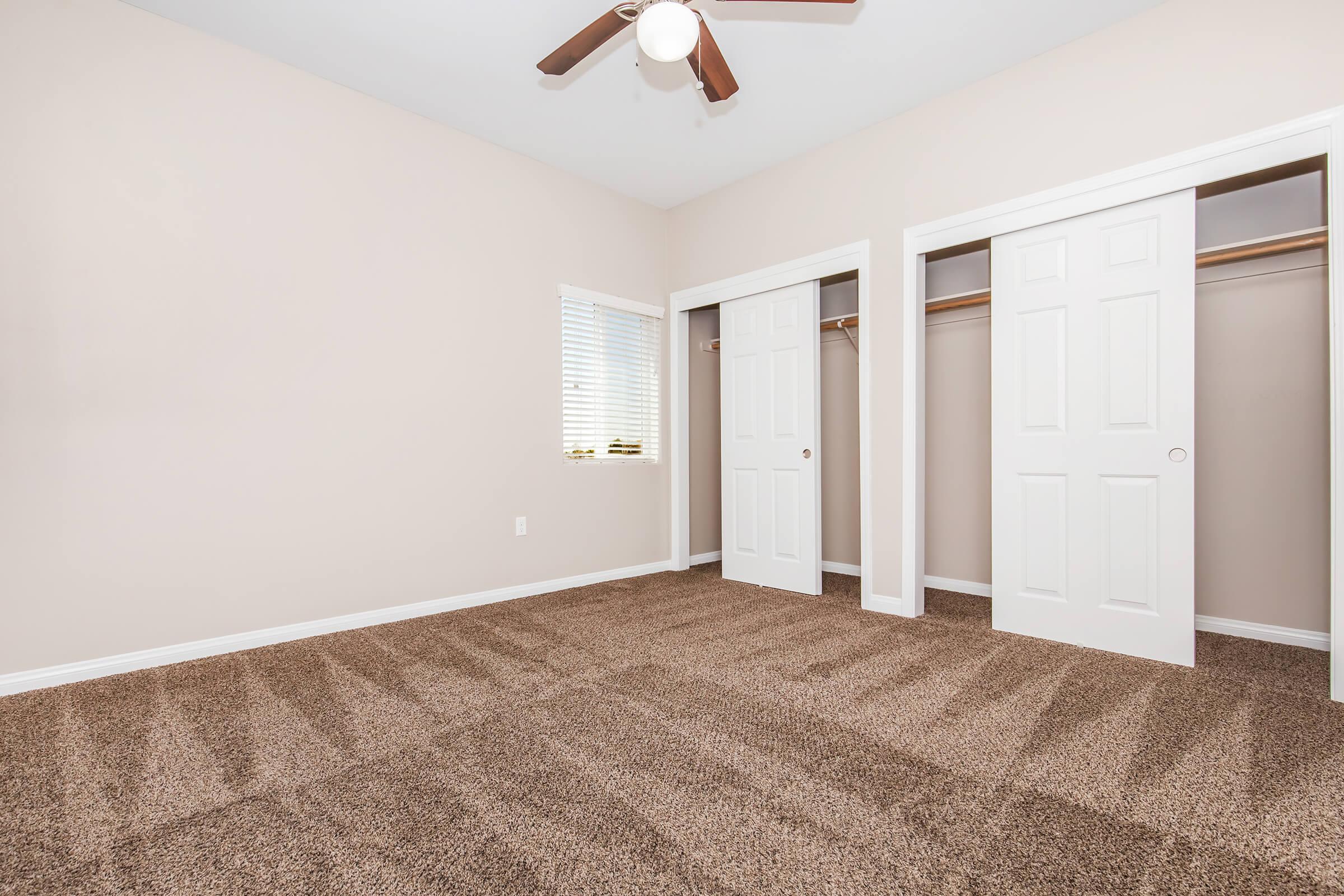 Empty room with light beige walls and a ceiling fan. There are two open closet doors on the right, revealing empty spaces. A small window on the left lets in natural light. The floor is covered in plush brown carpet, creating a cozy, inviting atmosphere.