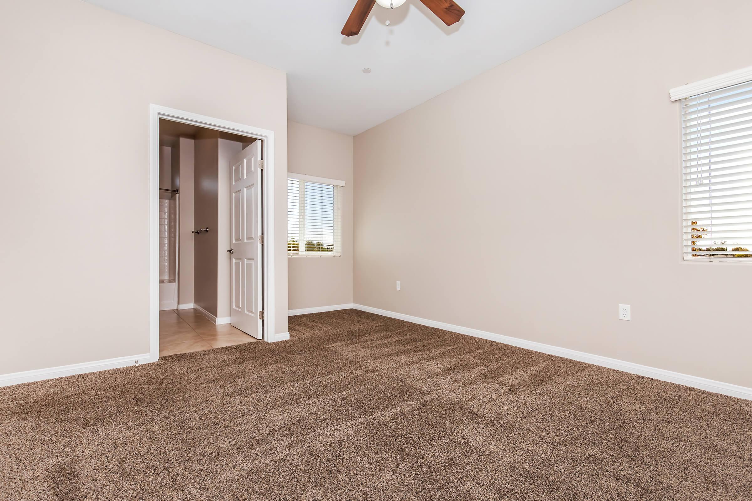 A spacious, empty room with light brown carpet and beige walls. There are two windows with white blinds allowing natural light in, and a ceiling fan is visible. A doorway leads to another room, likely a bathroom, in the background. The overall atmosphere is bright and clean, ready for furniture and personal touches.