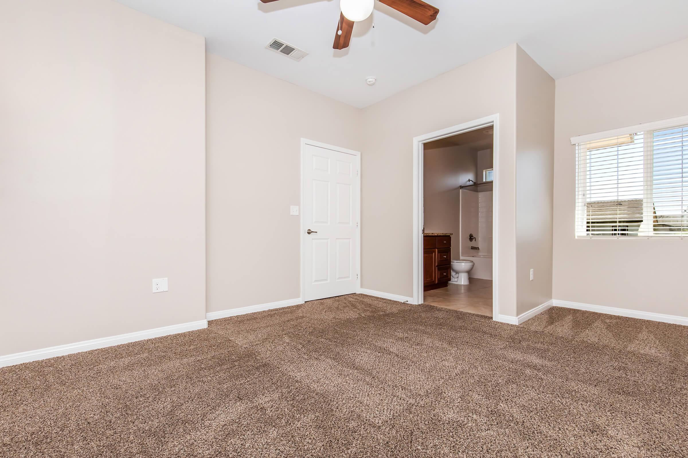 Empty room with light beige walls and brown carpet. A ceiling fan is mounted on the ceiling. To the right, there's an open door leading to a bathroom, visible behind a partially closed door, with a window letting in natural light. The room appears spacious and well-lit.