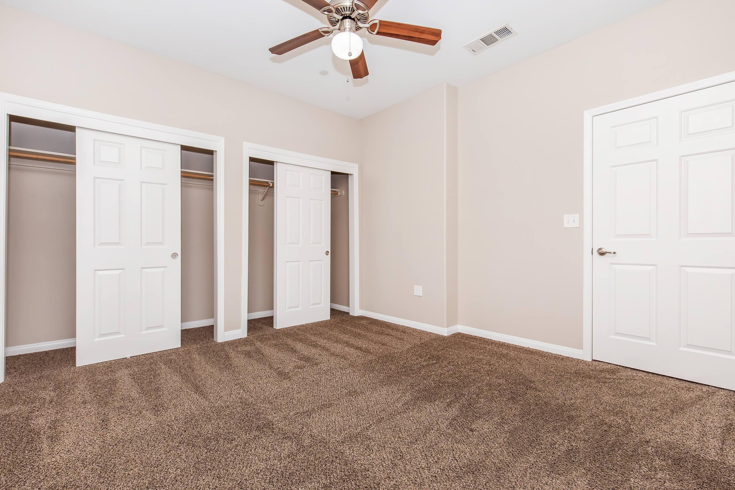 A spacious room with light beige walls and carpet flooring, featuring two closed closet doors on the left and a single door on the right. A ceiling fan with wooden blades is mounted above the room, providing a bright and airy feel to the space.