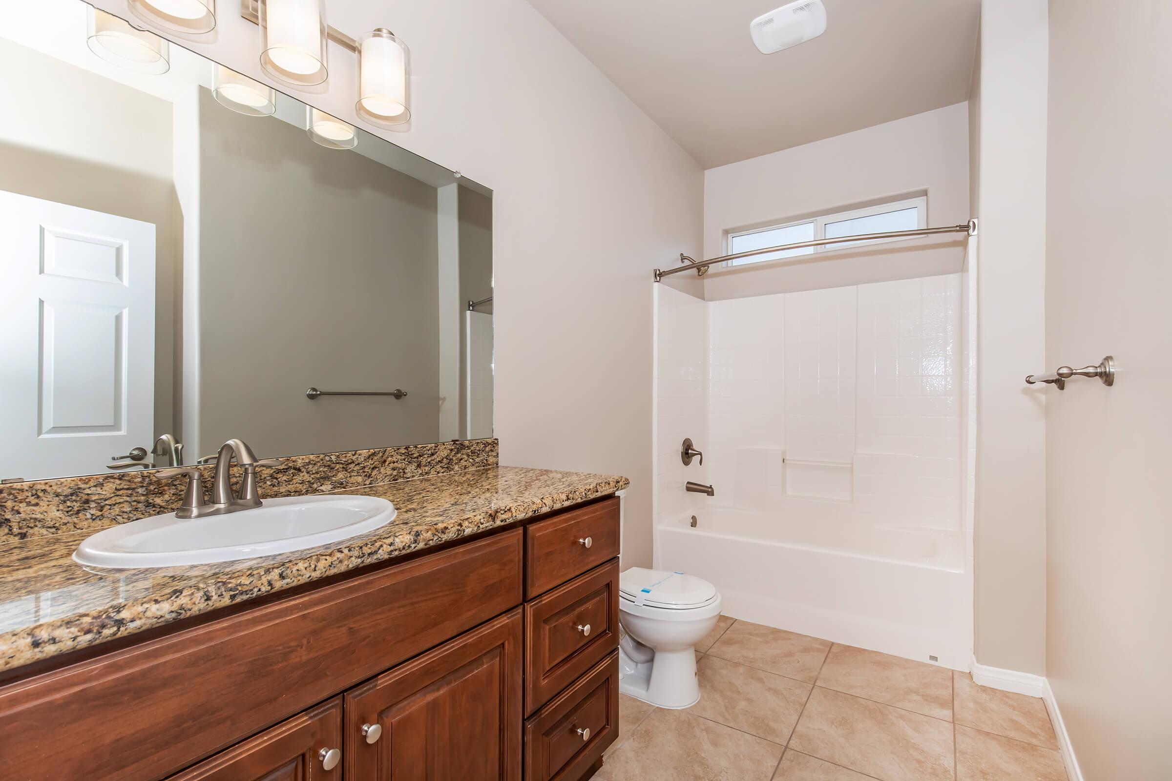 A modern bathroom featuring a granite countertop vanity with a white sink and mirror above. There is a toilet and a shower-tub combo with a shower curtain. The walls are painted a neutral color, and the floor is tiled. Natural light enters through a window above the tub.
