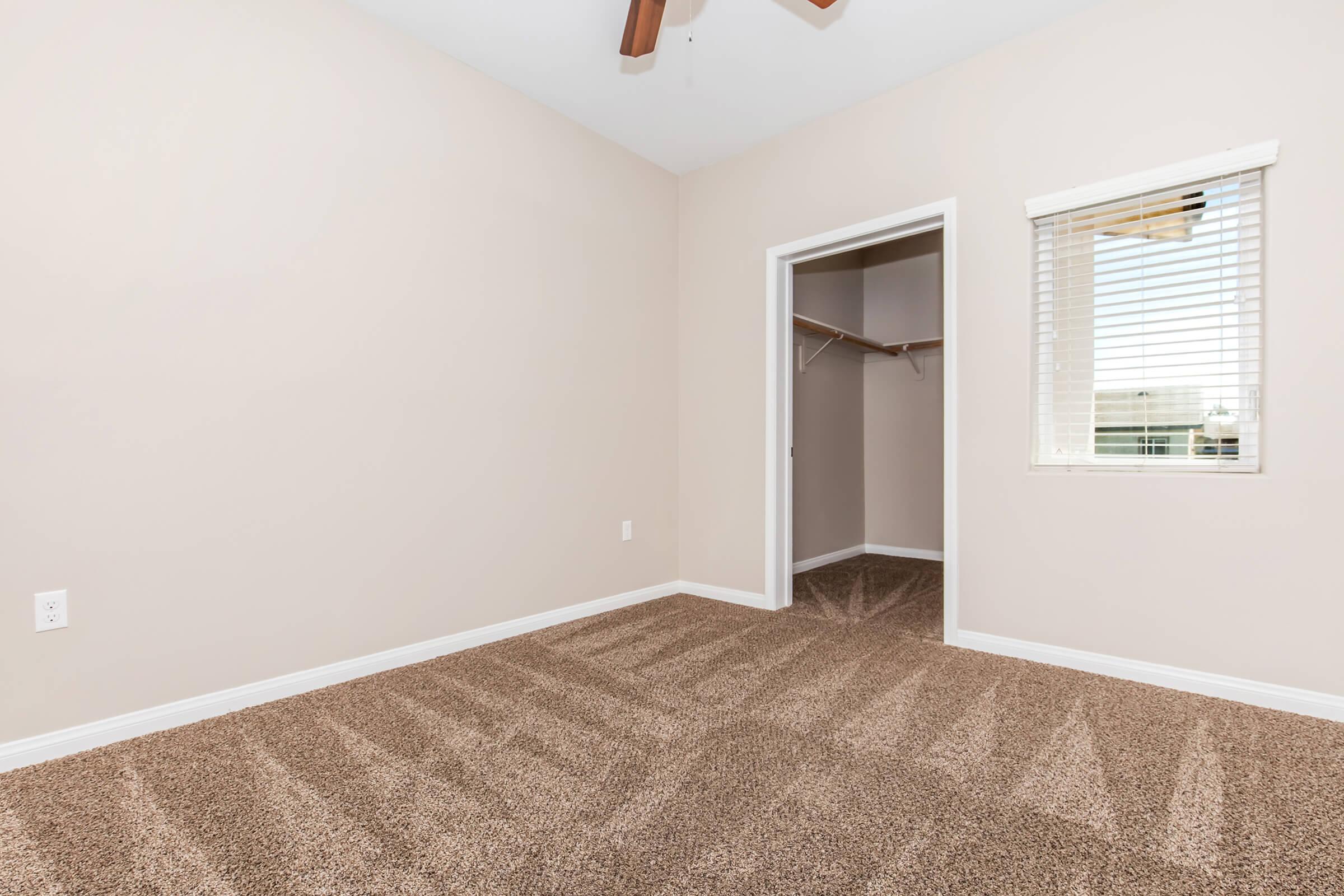 A light-colored bedroom featuring beige walls and carpeting. There is a ceiling fan with wooden blades, a small window with white blinds, and a closet with an open door. The room is empty, providing a blank canvas for decorating or furnishing.