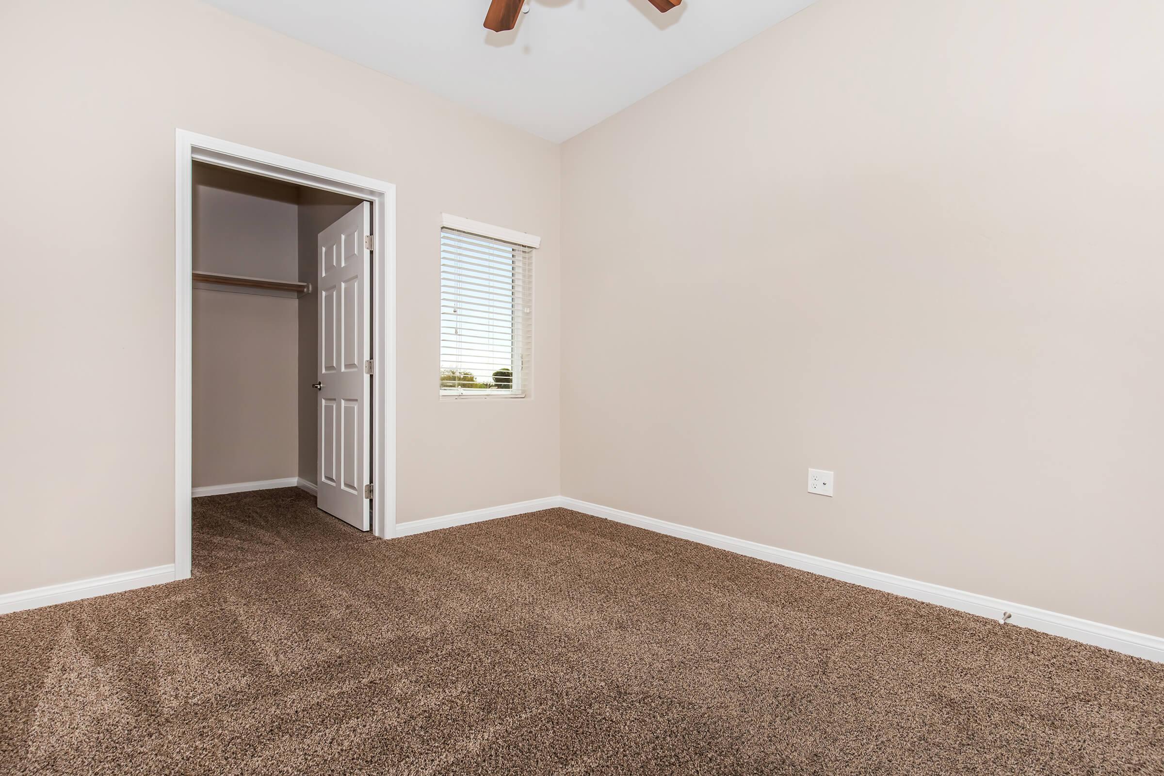 A well-lit, empty room featuring beige walls and new carpeting. There is a closet with a door on the left and a window with blinds on the right, allowing natural light to enter. The space appears clean and ready for furnishing, with a ceiling fan visible above.