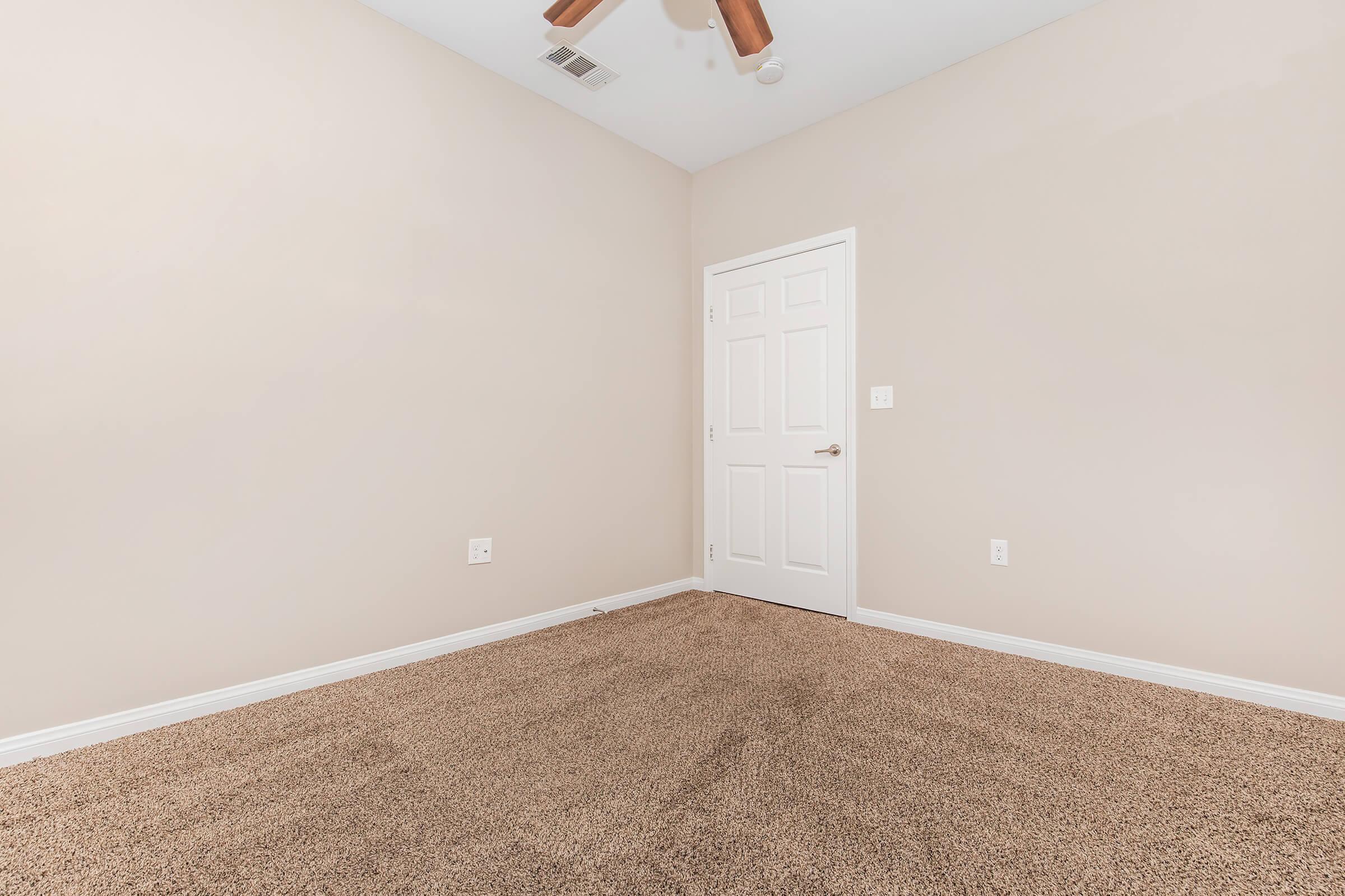 A minimalistic empty room with light beige walls and a carpeted floor. The ceiling features a ceiling fan, and there is a closed white door on the right. The space appears clean and ready for furnishing, with no visible decorations or furniture.