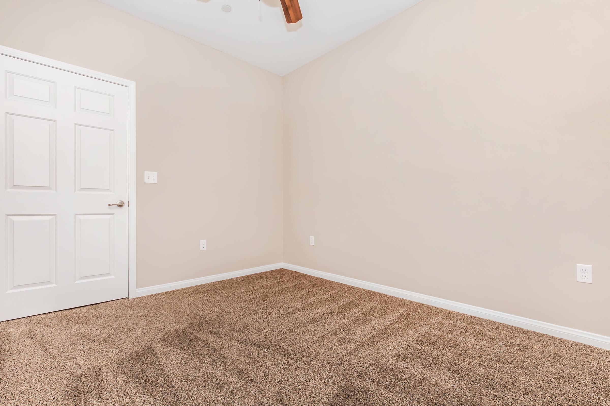 A vacant room with light beige walls and a ceiling fan. The floor is covered with tan carpet. There is a white door on the left and a single electrical outlet on the right wall. The room has a clean, minimalistic look with no furniture or decorations.