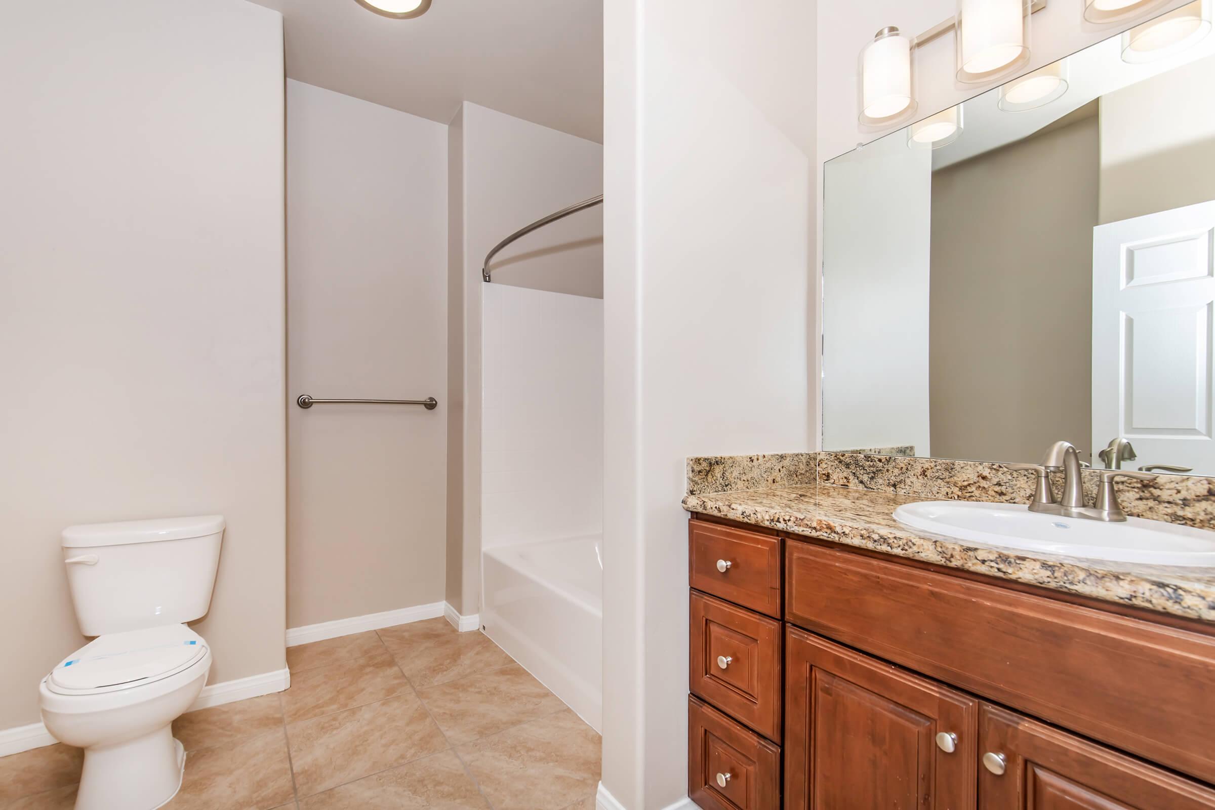 A modern bathroom featuring a white toilet, a shower with a curved curtain rod, and a granite countertop with a sink. The walls are painted a light color, and there is a large mirror above the sink. The flooring is tiled with a beige pattern.