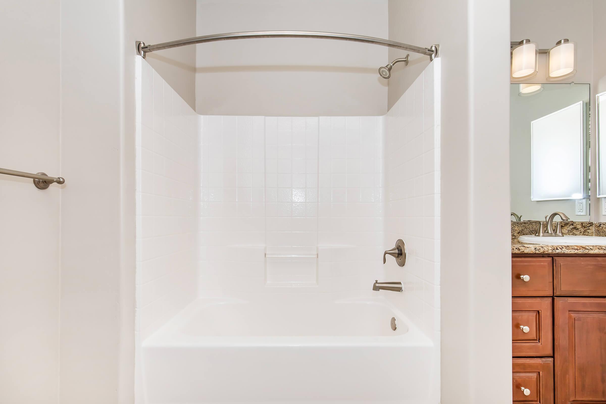 A clean, modern bathroom featuring a white tiled bathtub with an arch shower curtain rod. The tub has a chrome faucet and a small shelf in the wall. To the right, there is a wooden vanity with a granite countertop and a mirror above it. The walls are painted in a light color, creating a bright and airy atmosphere.