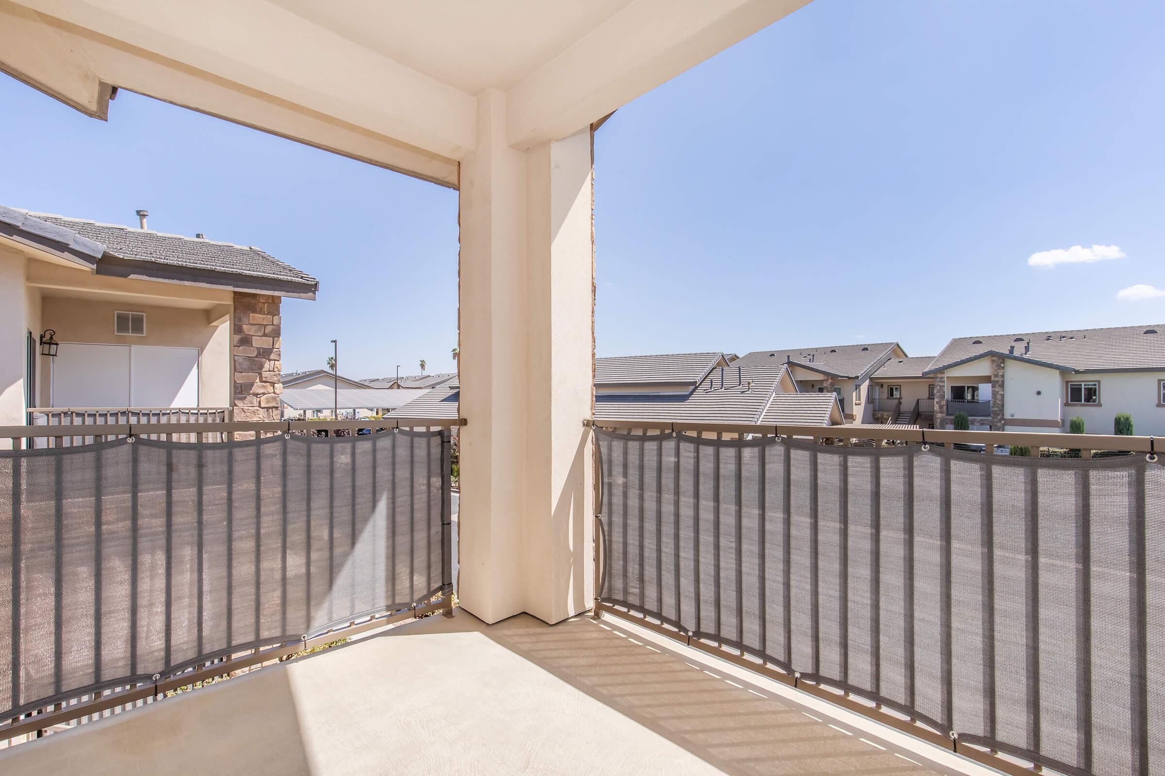 A view of a patio or balcony with a metal railing, featuring flooring and a clear blue sky in the background. Nearby buildings are visible, highlighting a residential area.