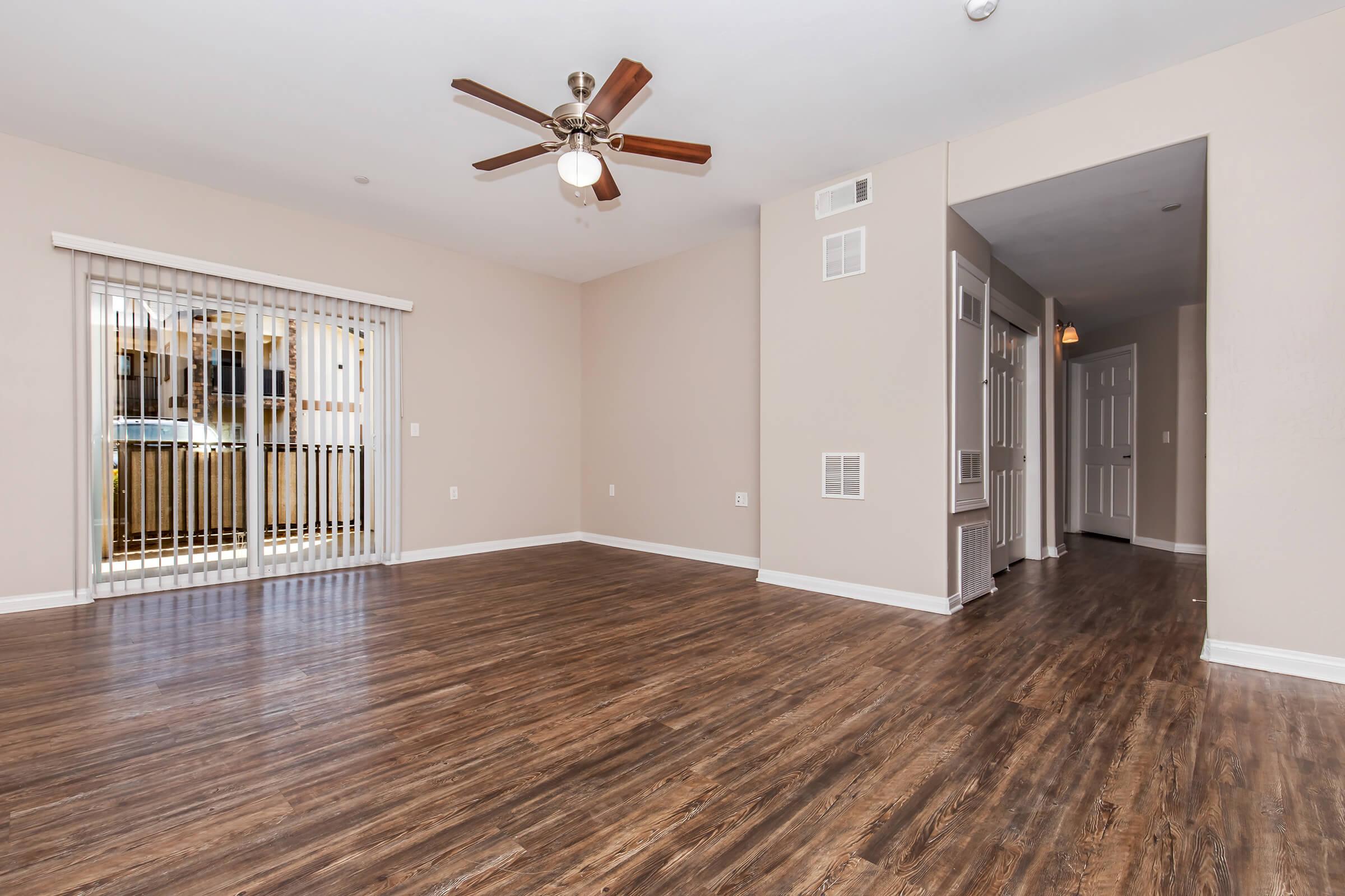 A spacious living room featuring a ceiling fan, hardwood-style flooring, light beige walls, and large sliding glass doors that open to a balcony. A hallway to the right leads to additional rooms, while a small heating unit is visible on the wall. The room is bright and inviting with ample natural light.