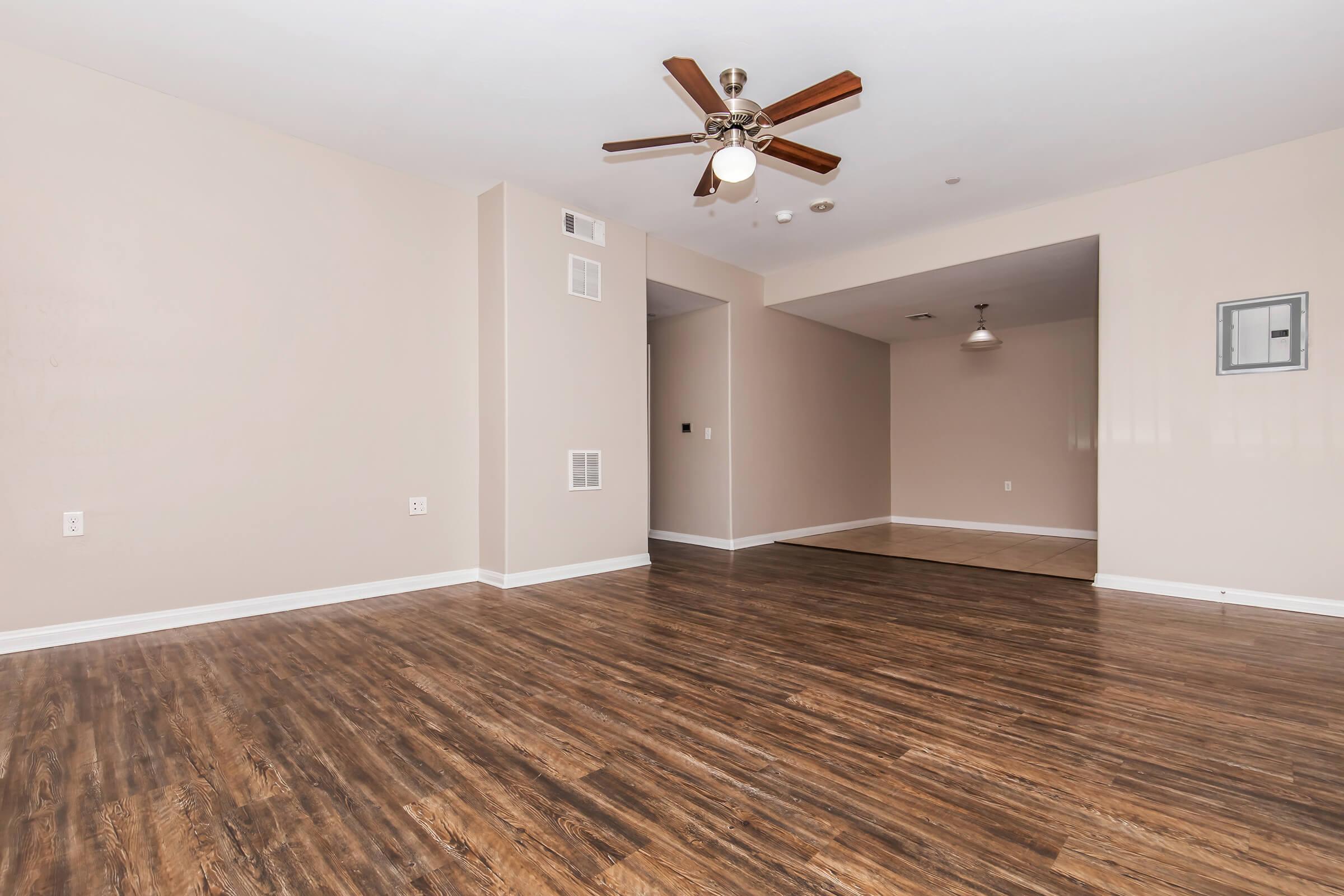 A spacious, empty living room with hardwood-style flooring, painted light beige walls, and a ceiling fan. There are no furnishings, creating an open and airy feel. A doorway leads to another room, and a light fixture hangs from the ceiling. The area is well-lit with natural light.