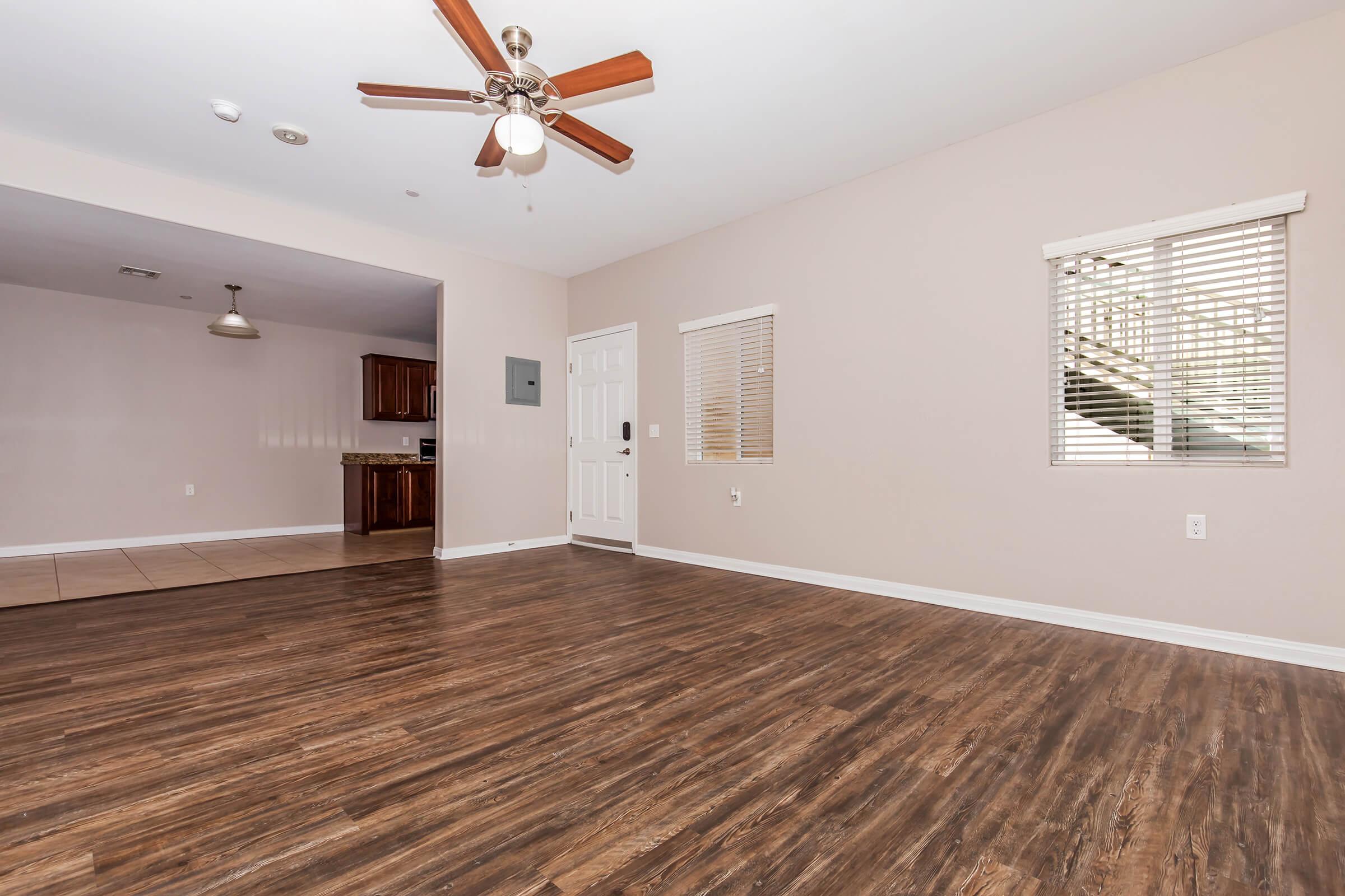 A spacious living area with hardwood floors, a ceiling fan, and neutral-colored walls. Windows allow natural light, and a doorway leads to a kitchen area with wooden cabinets visible in the background. The overall space is open and inviting, suitable for various interior design options.
