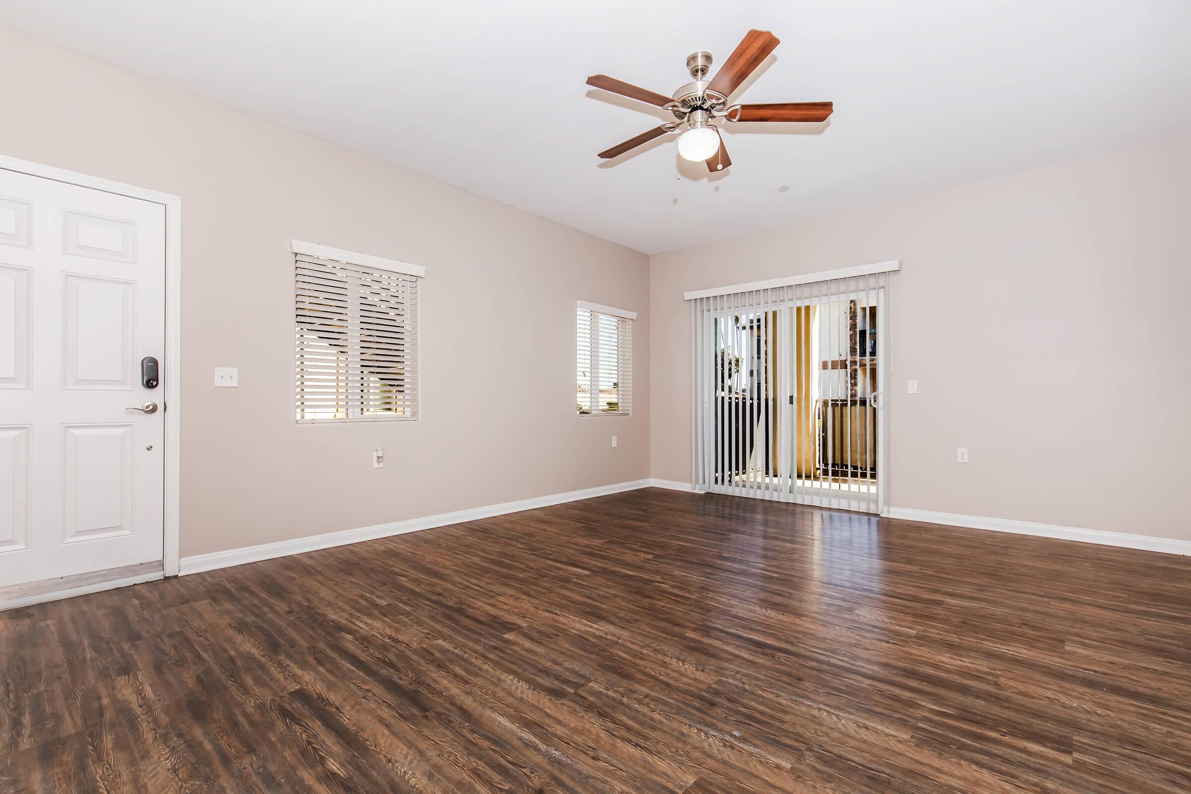 An empty room with light-colored walls and wooden flooring. It features a ceiling fan with wooden blades, a door on the left, and two windows with blinds. A sliding glass door leads to an outdoor area, allowing natural light to enter the space. The room has a spacious, airy feel.