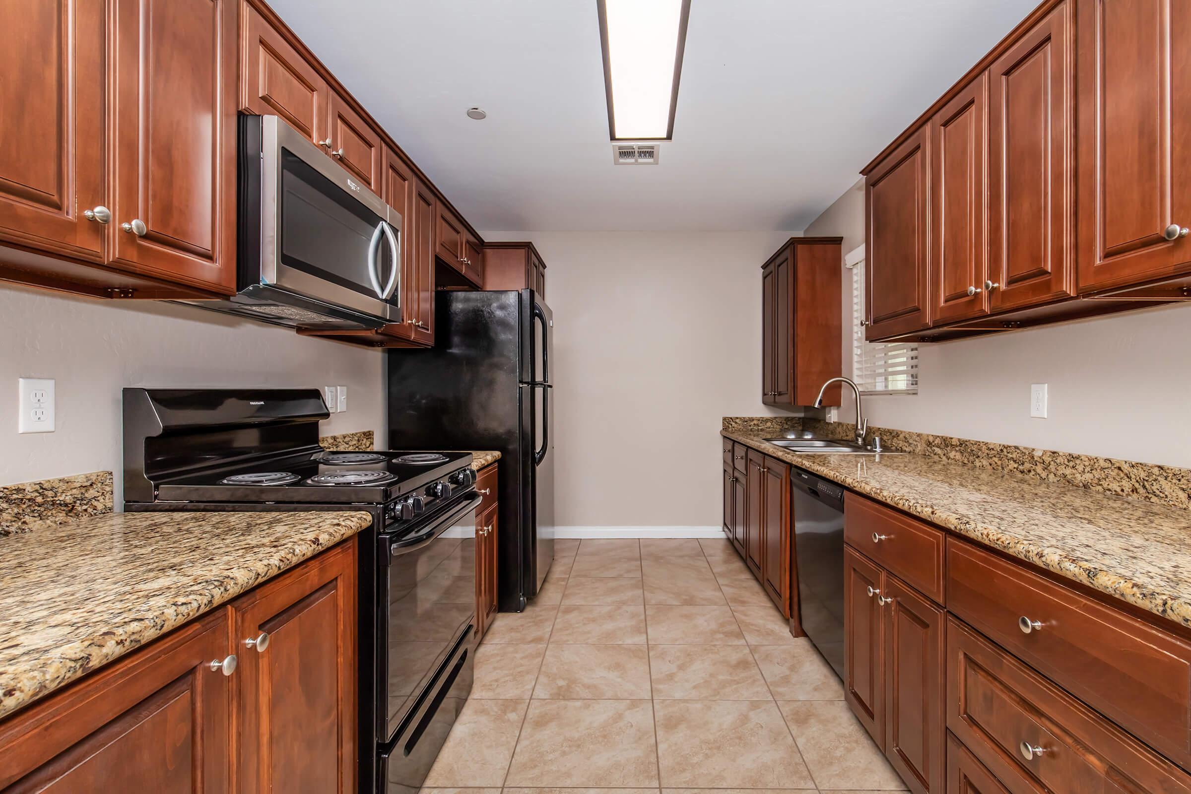 Modern kitchen featuring dark wooden cabinets, stainless steel appliances including a microwave and a gas stove, and granite countertops. The room has beige walls and tiled flooring, with a single sink and window, creating a bright and functional cooking space.
