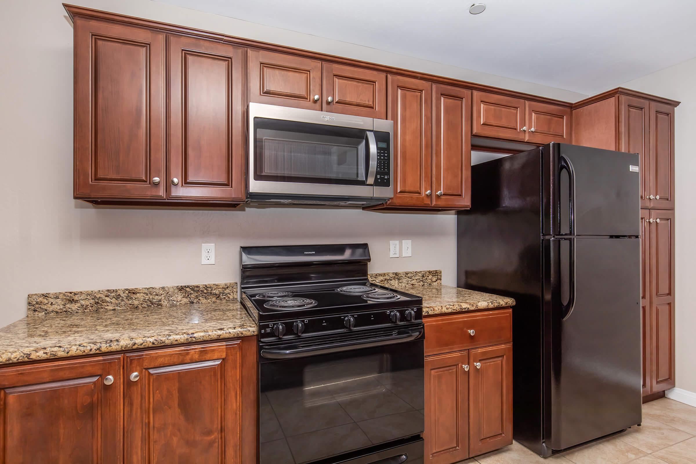 Modern kitchen featuring wooden cabinets, a black stove, and a microwave above it. A black refrigerator is positioned next to the cabinets, and the countertops are made of granite. The walls are painted a neutral color, creating a warm, inviting atmosphere.