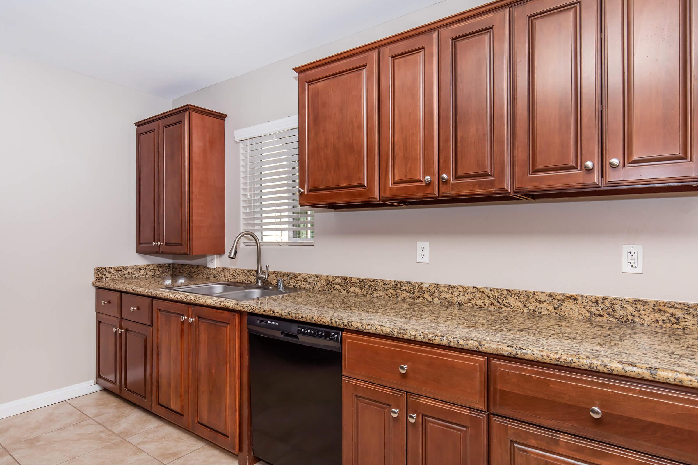A modern kitchen featuring dark wood cabinets, a granite countertop, a sink with a single faucet, and a black dishwasher. There is also a window with blinds above the sink, and beige tile flooring completes the look.
