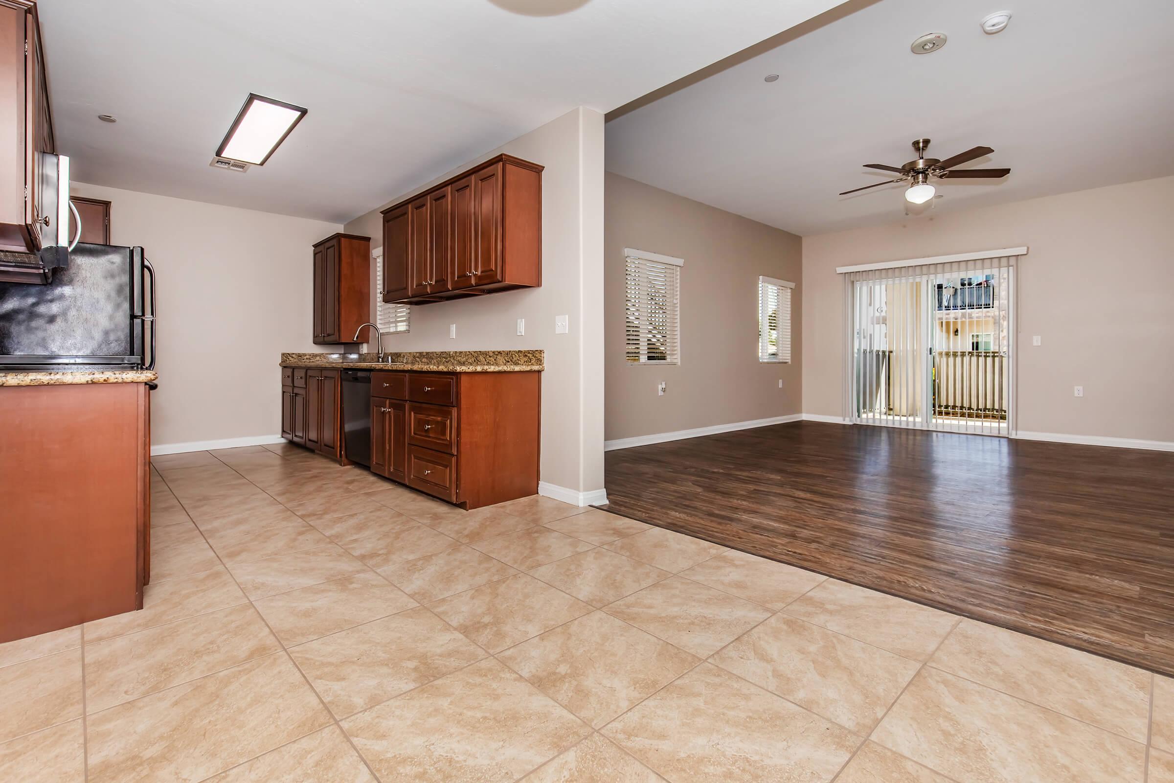 Spacious interior of an apartment featuring a kitchen with dark wood cabinets and granite countertops, adjacent to a living area with a ceiling fan and large windows. The floor is a mix of tiled and hardwood surfaces, with sliding doors leading to a balcony or outdoor area.