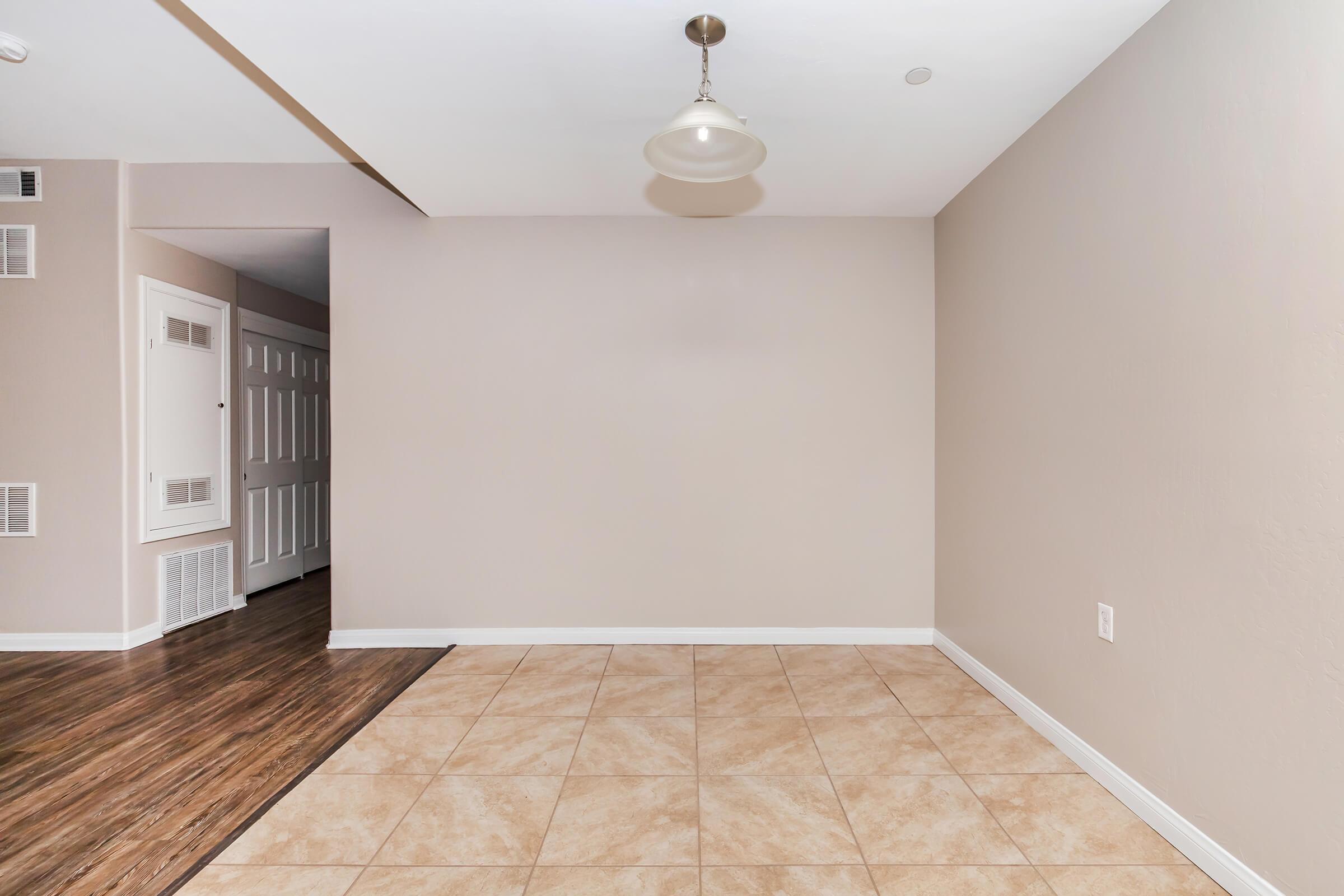 A spacious, empty room featuring beige walls and a light fixture. The flooring consists of tile and hardwood. There is a doorway leading to another area, and wall vents are visible. The room is well-lit, creating a clean and inviting atmosphere.