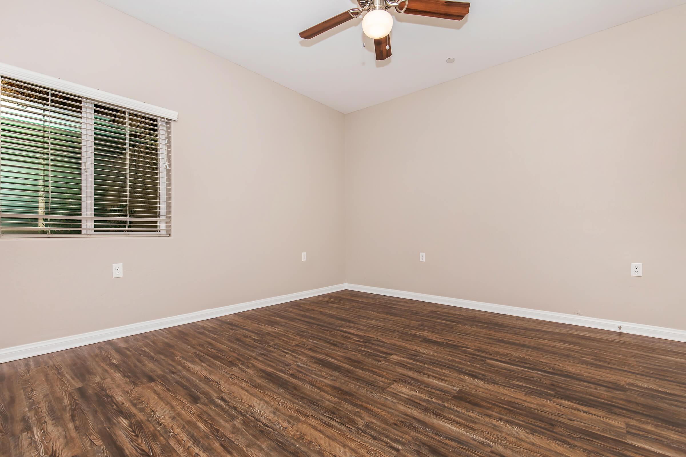 Empty room with light brown walls and a ceiling fan. The floor is wooden laminate, and there are windows with blinds allowing natural light to enter. The space is uncluttered and ready for furniture placement.