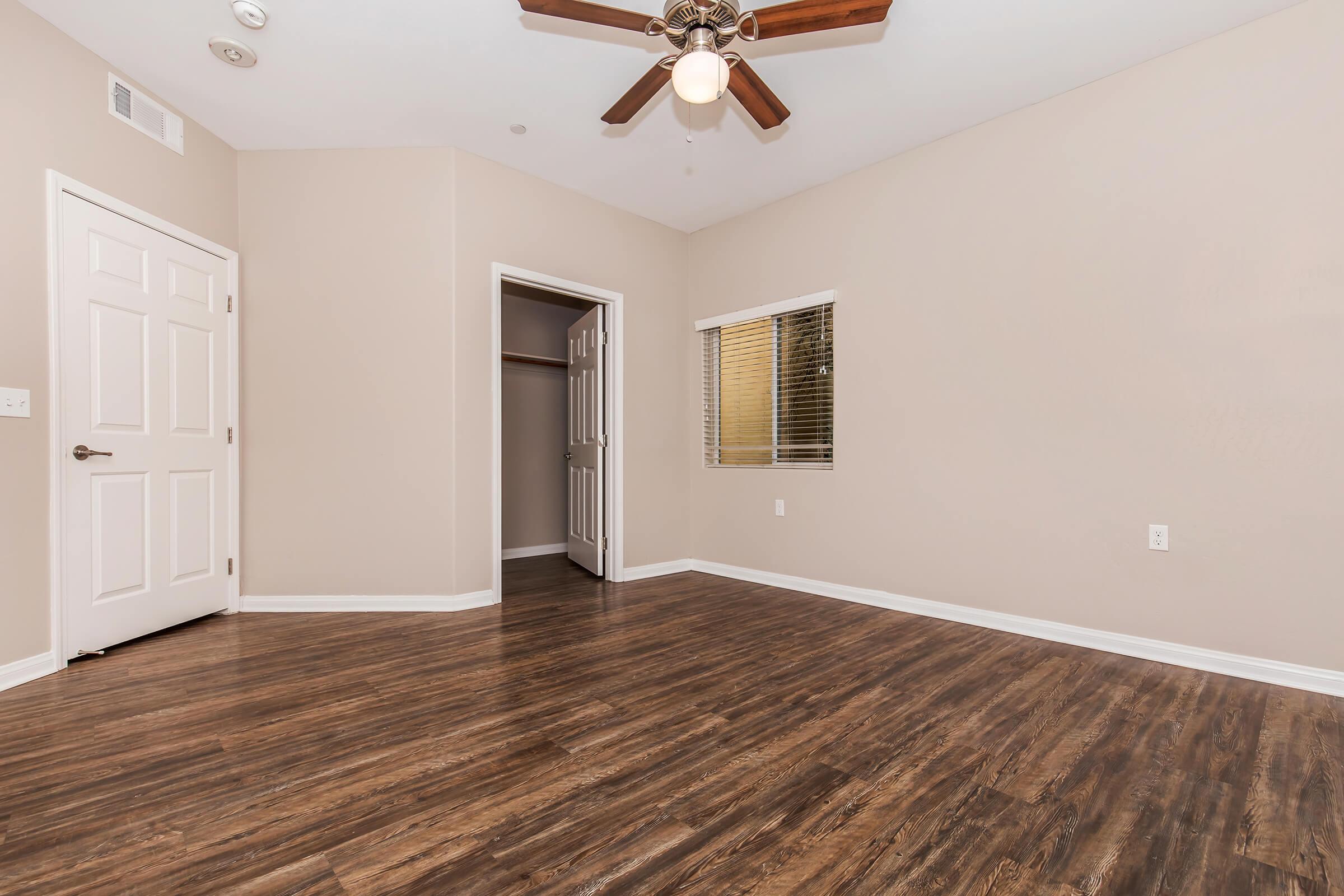 Interior of a room with light brown walls and hardwood flooring. The room features a ceiling fan with five blades, a window with blinds, and a door leading to a closet. The space is empty, creating a bright and airy atmosphere.