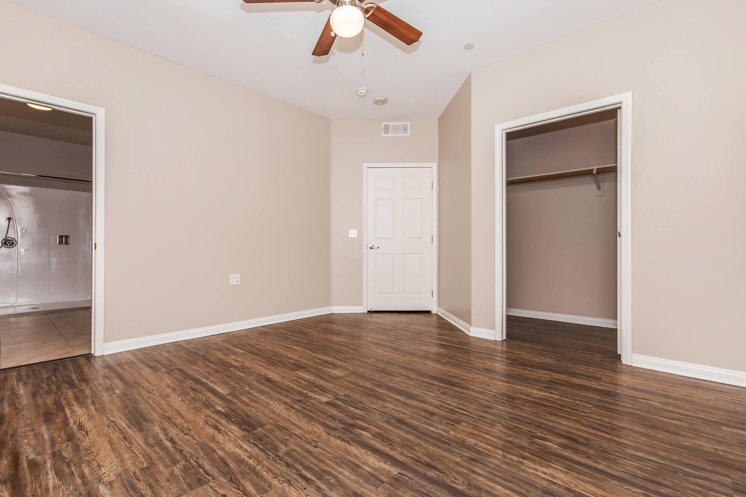 A spacious empty room with light beige walls and a ceiling fan. The flooring is a rich brown wood-like laminate. A door leads to a closet on the left, while another door is on the right. Natural light enters through the doorway, enhancing the bright and open feel of the space.