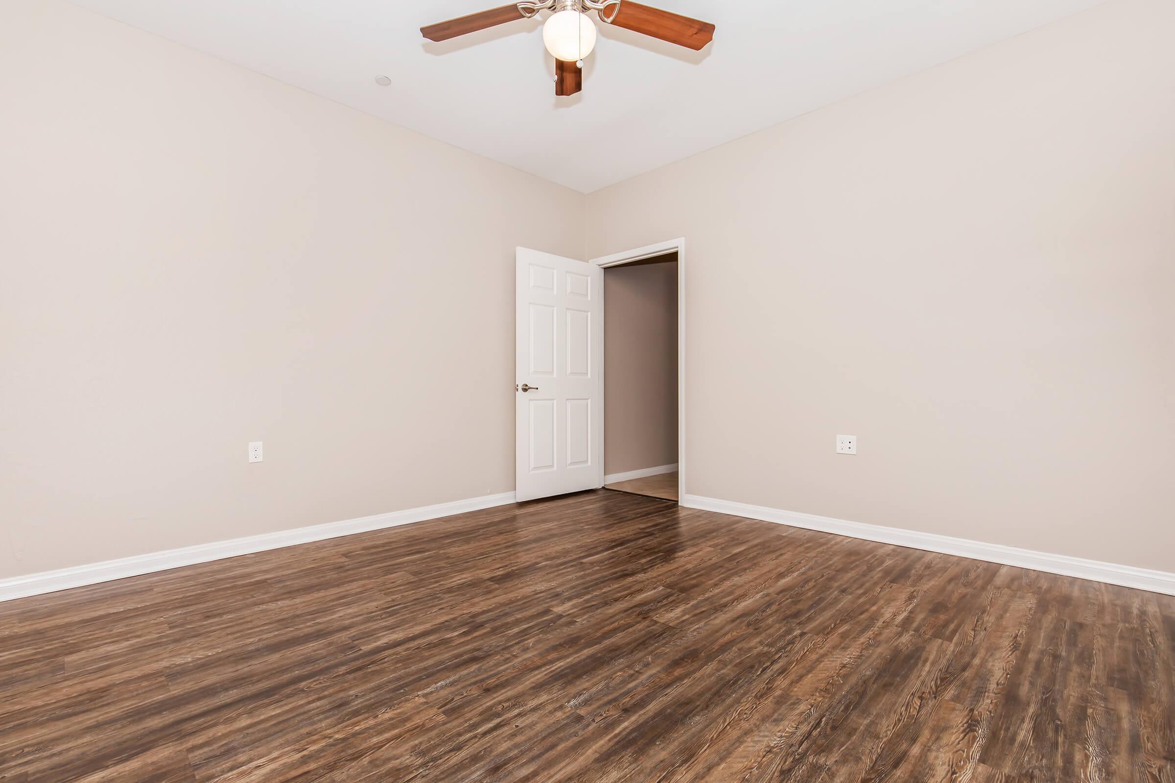 A spacious, empty room with light beige walls and a dark wood floor. A ceiling fan is mounted above, and there is a single white door on the left leading to another room. The overall atmosphere is bright and clean, suitable for a fresh start or personal decoration.
