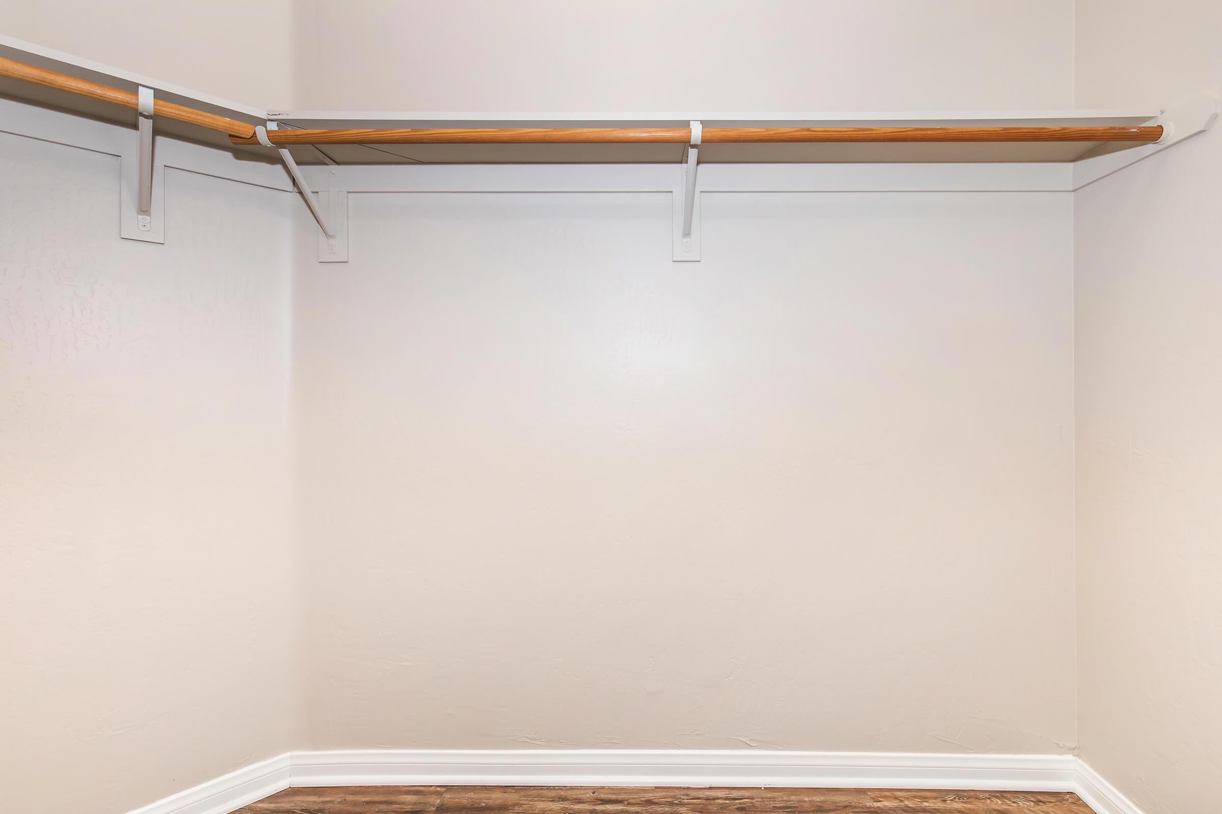 Empty closet with a wooden hanging rod and a light-colored wall. The floor is dark, possibly wood, and there are no items or shelves present. The space appears clean and minimalistic.