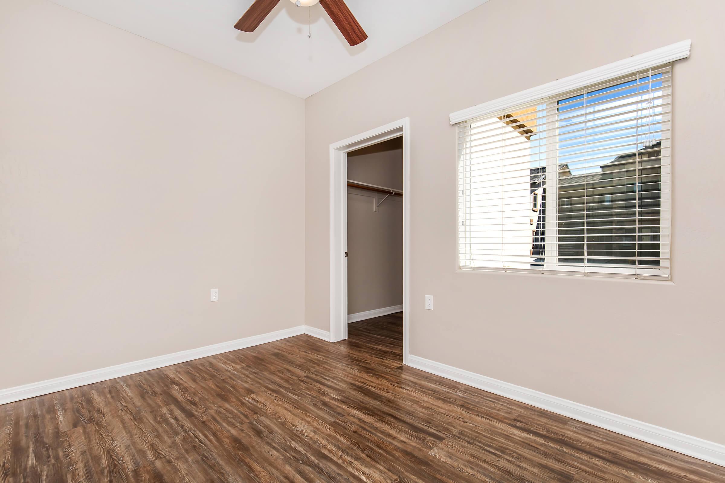 A clean, empty room with light brown walls and dark wooden flooring. There is a ceiling fan and a window with blinds that lets in natural light. A closet space is visible through an open door, and the overall atmosphere is bright and spacious, suitable for various potential uses.