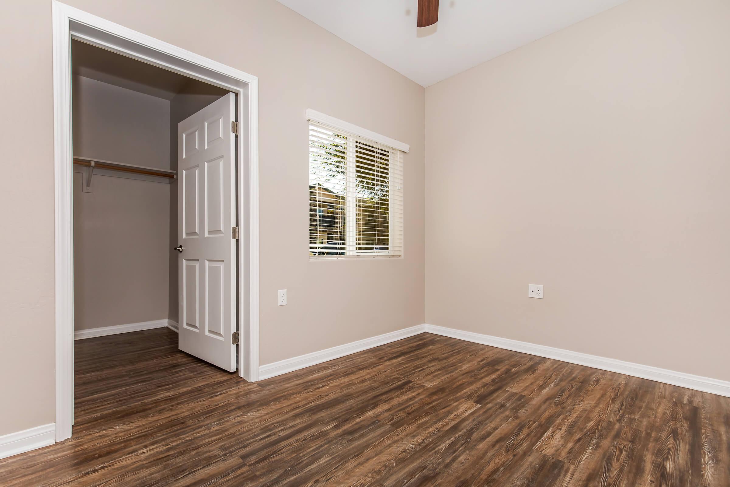 A clean, vacant room featuring light beige walls, wooden flooring, and a window with blinds. An open door leads to a closet on the left side, and there are no furnishings present, creating a spacious and bright atmosphere. The room is well-lit with natural light coming through the window.