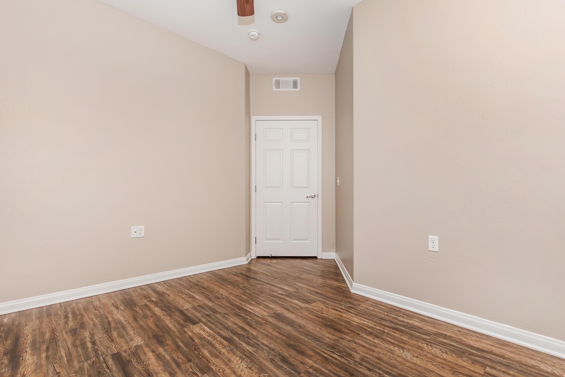 A small, empty room with light beige walls and brown laminate flooring. There is a white door on the left, a ceiling fan above, and no furniture or decorations. The overall atmosphere is clean and minimalistic.