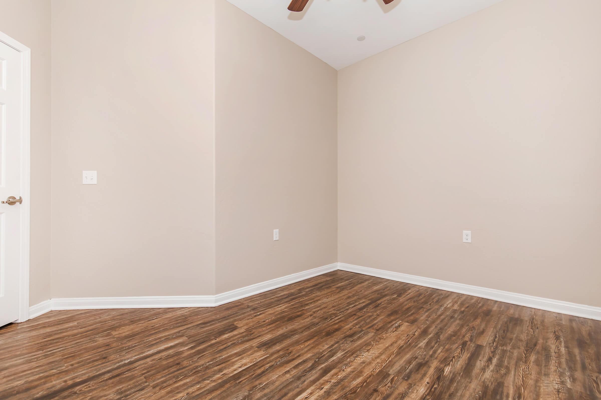 A spacious empty room with light beige walls and a wooden floor. There is a white door on the left, and the ceiling features a ceiling fan. The room is well-lit, creating a warm and inviting atmosphere.