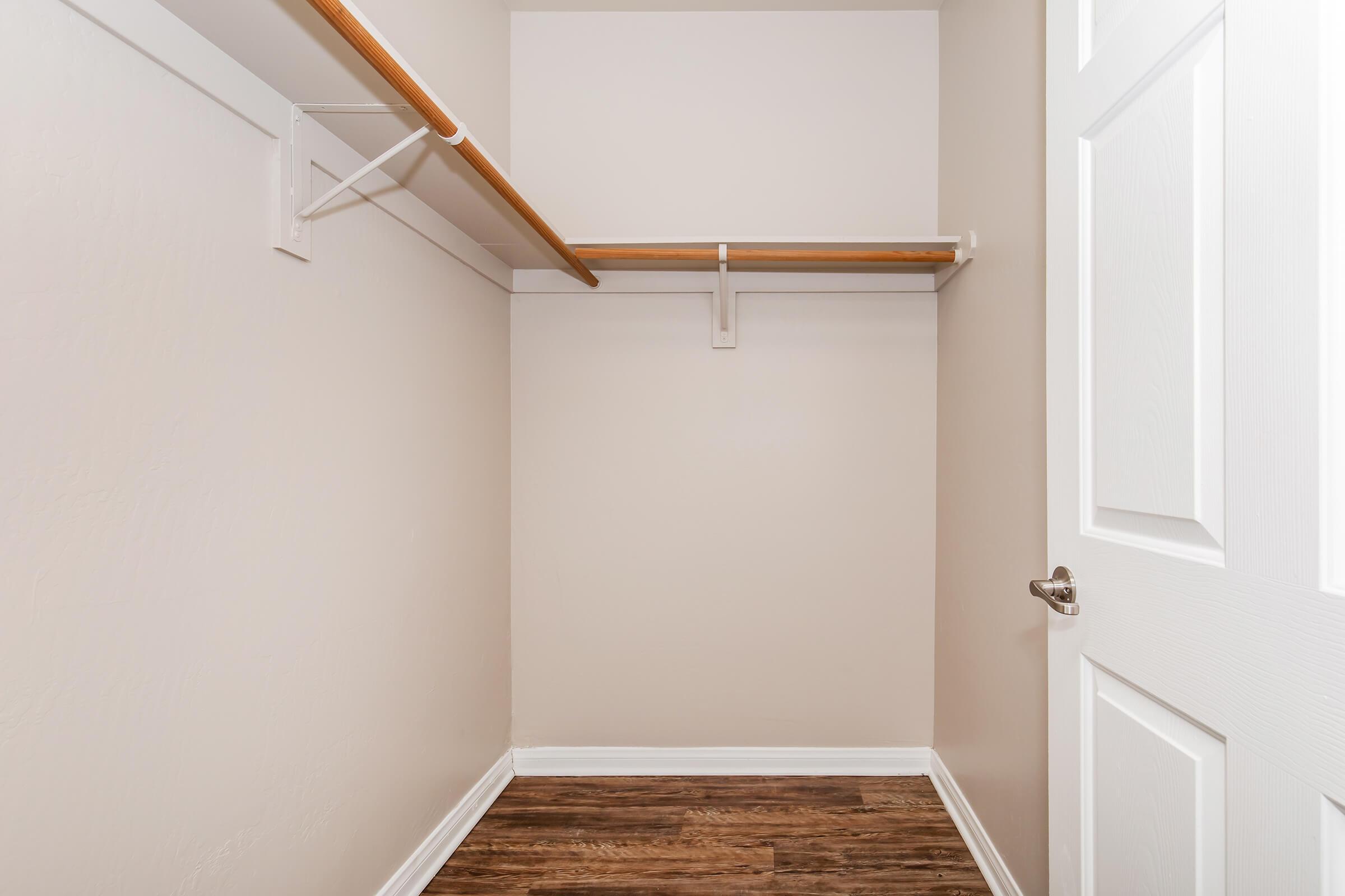 An empty closet with light beige walls and a wooden shelf at the top. The floor is made of dark wood, and there is a white door with a silver handle partially visible. The space appears clean and organized, suitable for storage.