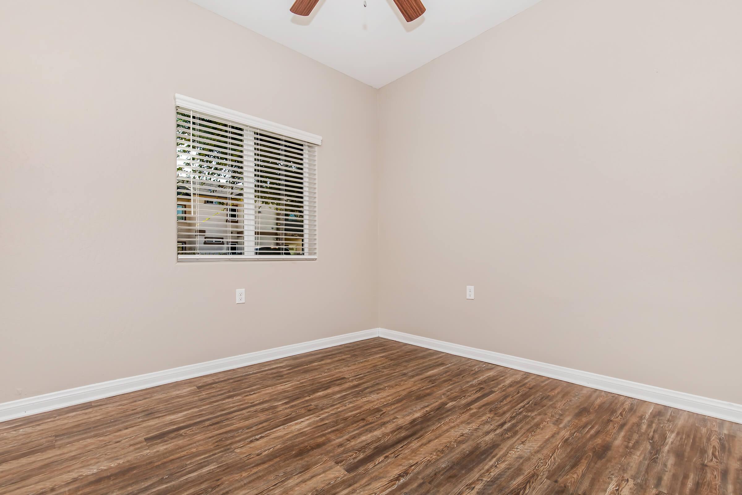 Empty room with light beige walls and wooden flooring. A ceiling fan is visible, and there is a window with white blinds allowing natural light to enter. The space appears clean and ready for decoration or furniture placement.