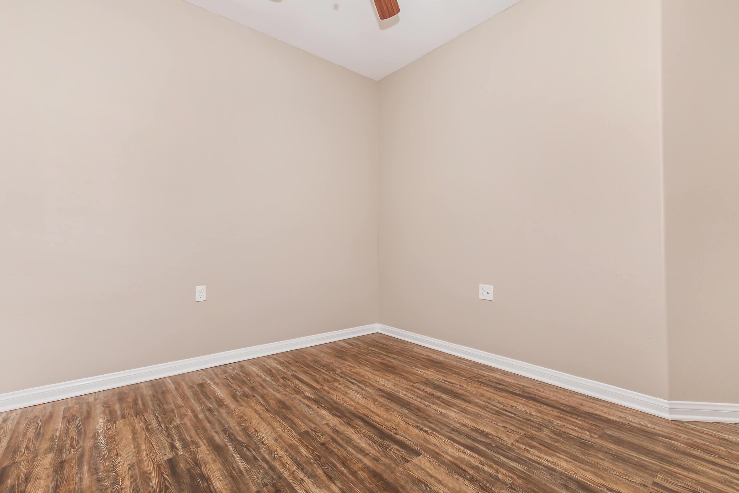 Empty room with light beige walls and a wooden floor. The room has a corner without furniture, featuring a ceiling fan. There are two electrical outlets visible on the walls. The overall ambiance is bright and minimalistic, providing a blank canvas for decoration or furniture placement.