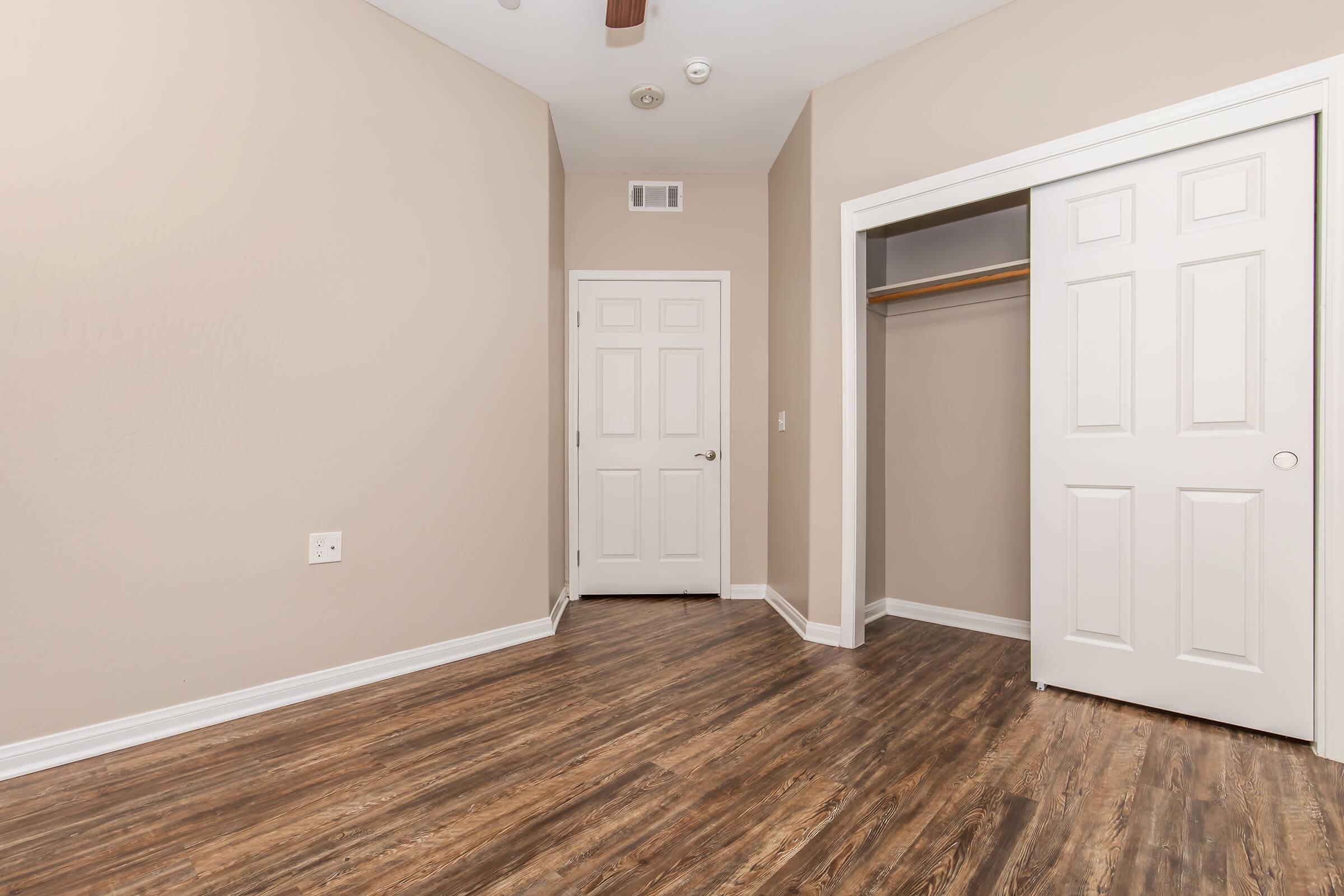 A clean, empty room featuring light brown walls and a light wood floor. There's a closed white closet door on the right, a plain white door visible in the background, and a ceiling fan. Natural light may be entering from an unseen window, enhancing the spacious feel of the area.