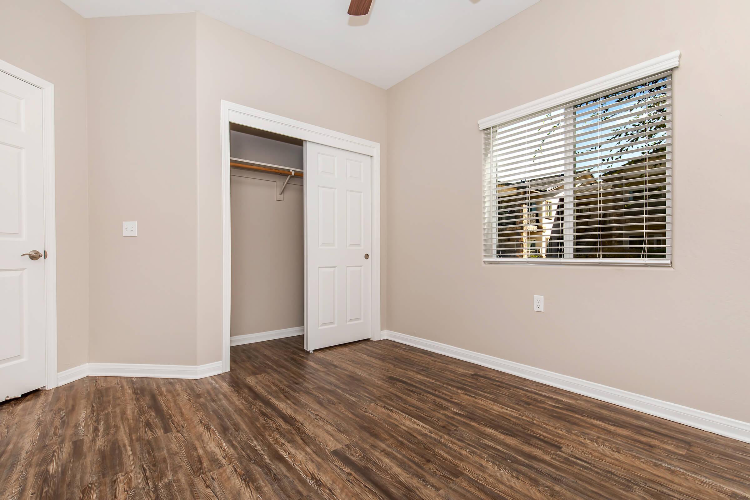 A light-colored room featuring laminate wood flooring, white walls, and a ceiling fan. There is a closed white door on the left leading to a closet, a window with blinds allowing natural light, and another wall with a power outlet. The space appears clean and uncluttered, suitable for various uses.