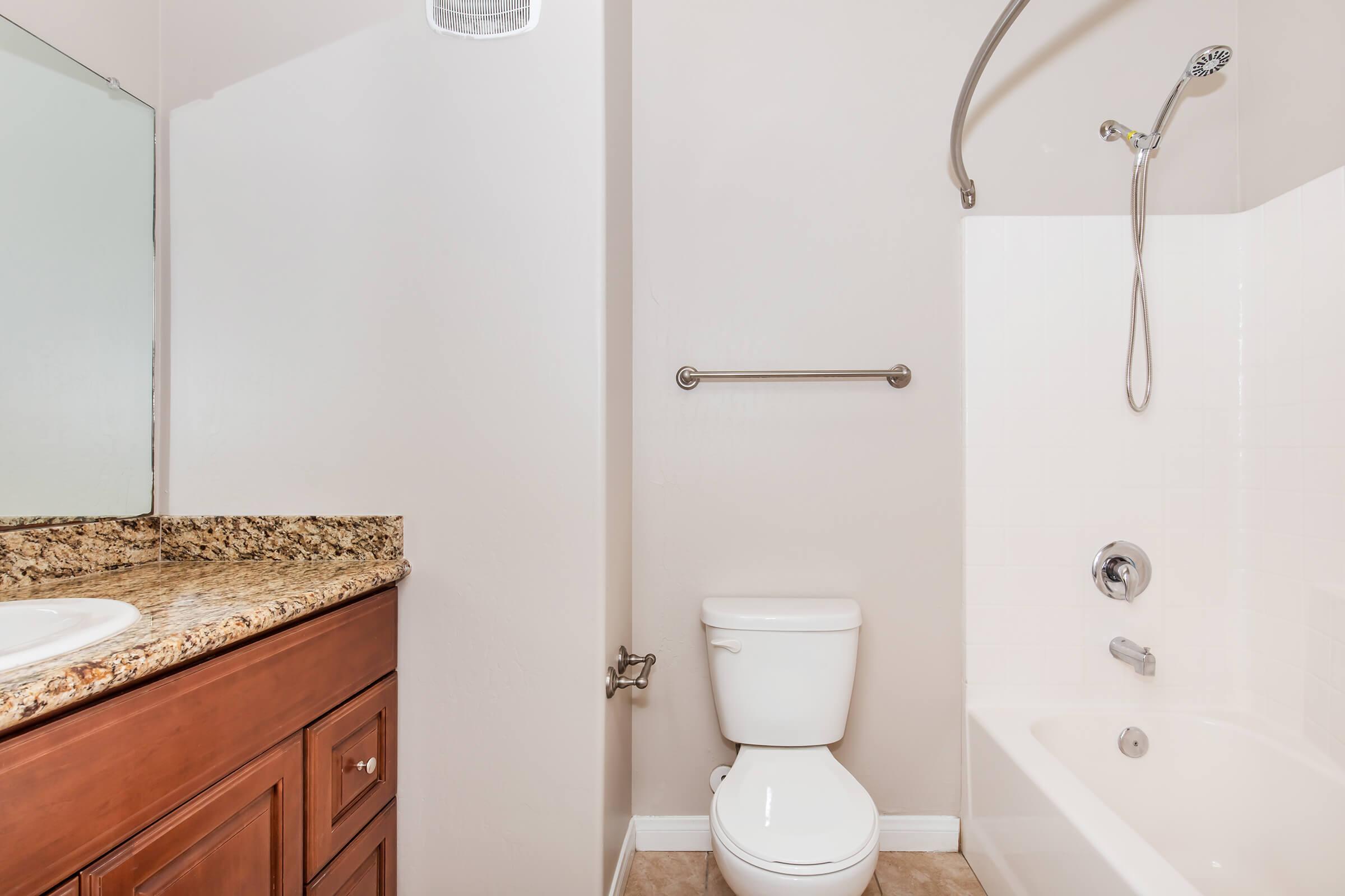 A bathroom featuring a shower and bathtub combination, white tiles, and a light-colored wall. A wooden vanity with a granite countertop is visible, along with a modern toilet and metal grab bar. The space is well-lit and clean, creating a minimalist aesthetic.
