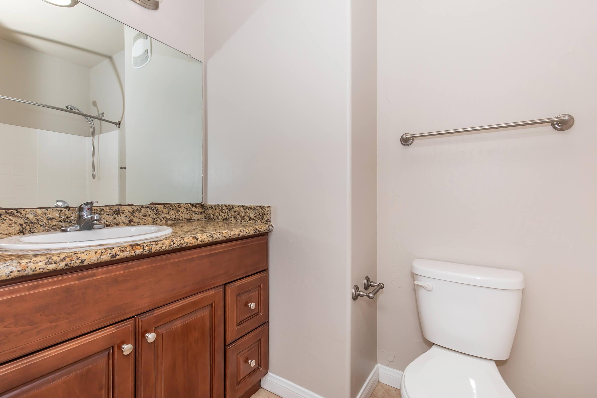 A clean bathroom featuring a wooden vanity with a granite countertop, a mirror above the sink, a wall-mounted towel bar, and a white toilet. The space is well-lit with beige walls, and a shower with a glass door is visible in the background.